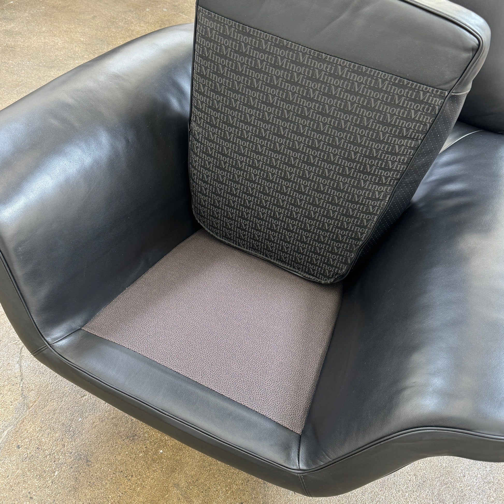 A Minotti Blake Lounge Chair & Ottoman by Minotti in black leather with chrome swivel bases, positioned on a bare concrete floor against a plain light-colored wall.