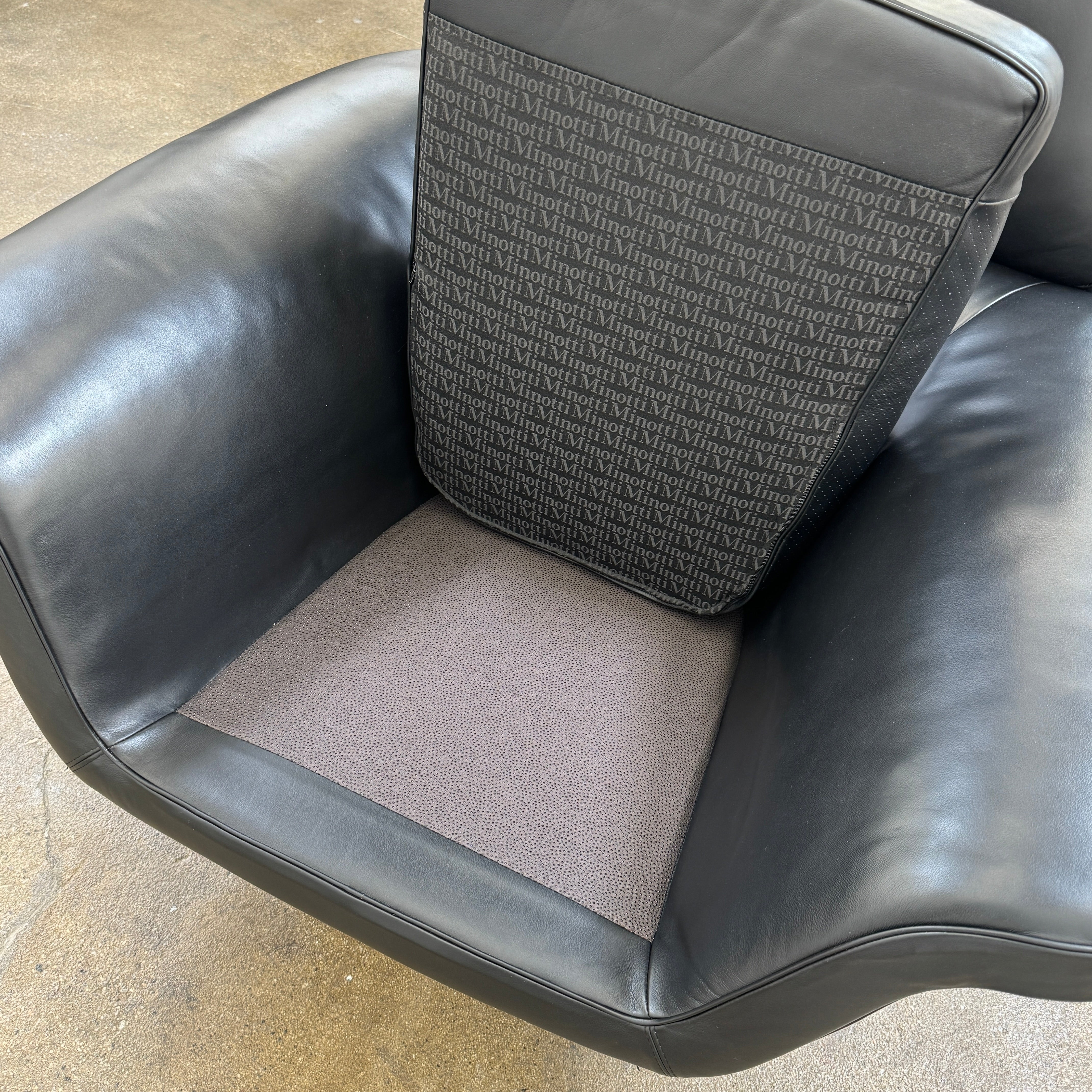 A Minotti Blake Lounge Chair & Ottoman by Minotti in black leather with chrome swivel bases, positioned on a bare concrete floor against a plain light-colored wall.