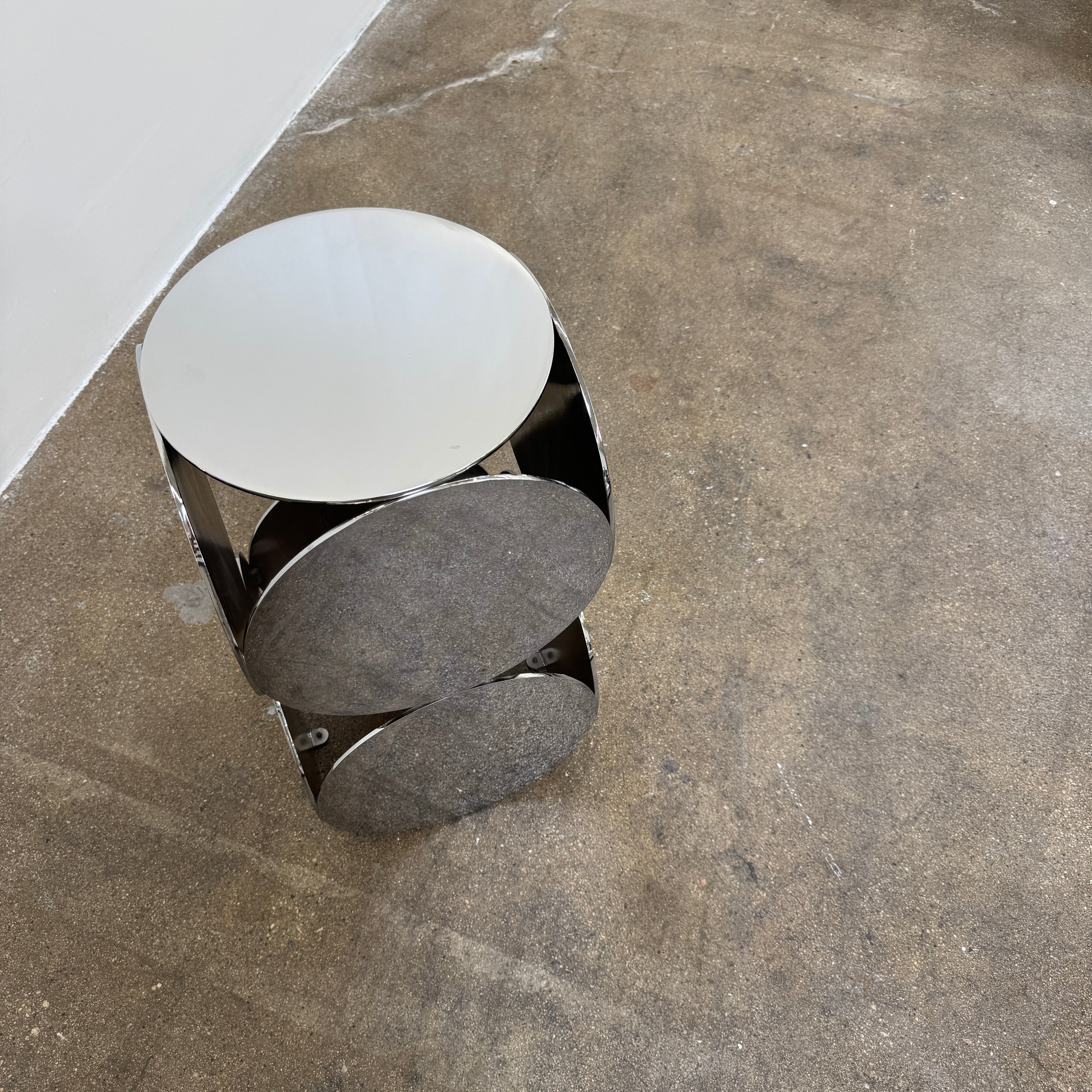 The Minotti Still Accent Table by Minotti features four large, circular, reflective metal discs stacked vertically for minimal elegance, shown on a concrete floor against a white wall.