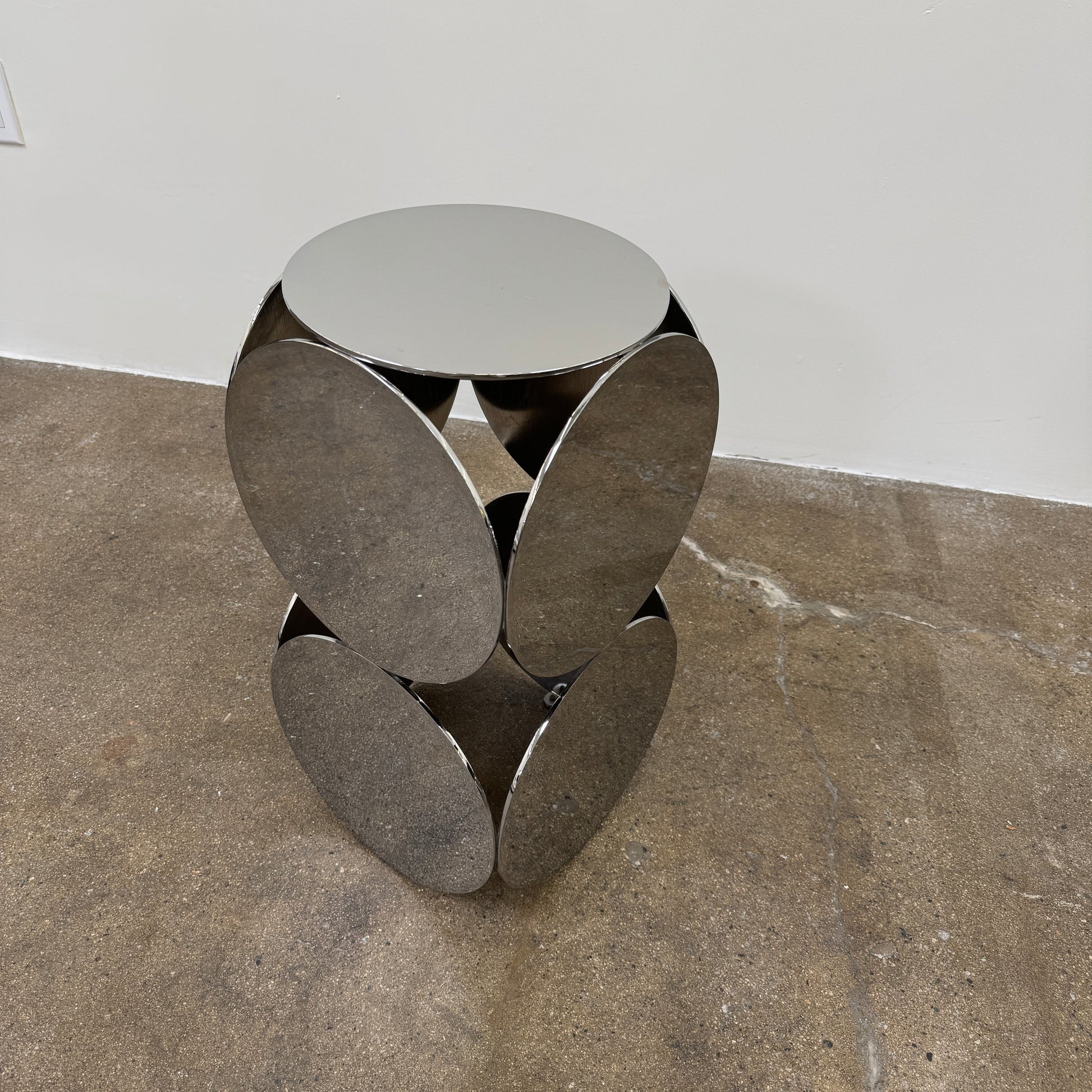 The Minotti Still Accent Table by Minotti features four large, circular, reflective metal discs stacked vertically for minimal elegance, shown on a concrete floor against a white wall.