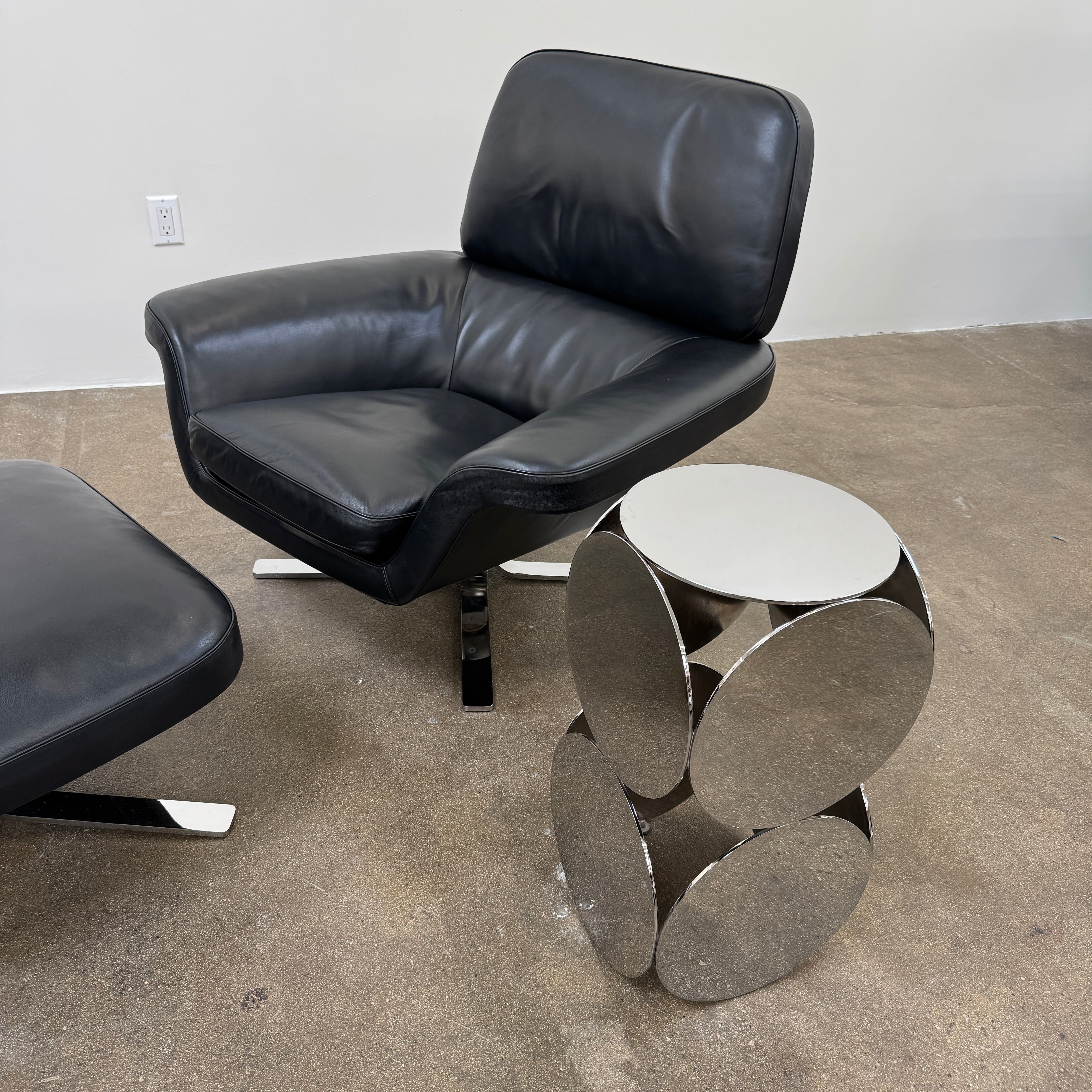 The Minotti Still Accent Table by Minotti features four large, circular, reflective metal discs stacked vertically for minimal elegance, shown on a concrete floor against a white wall.