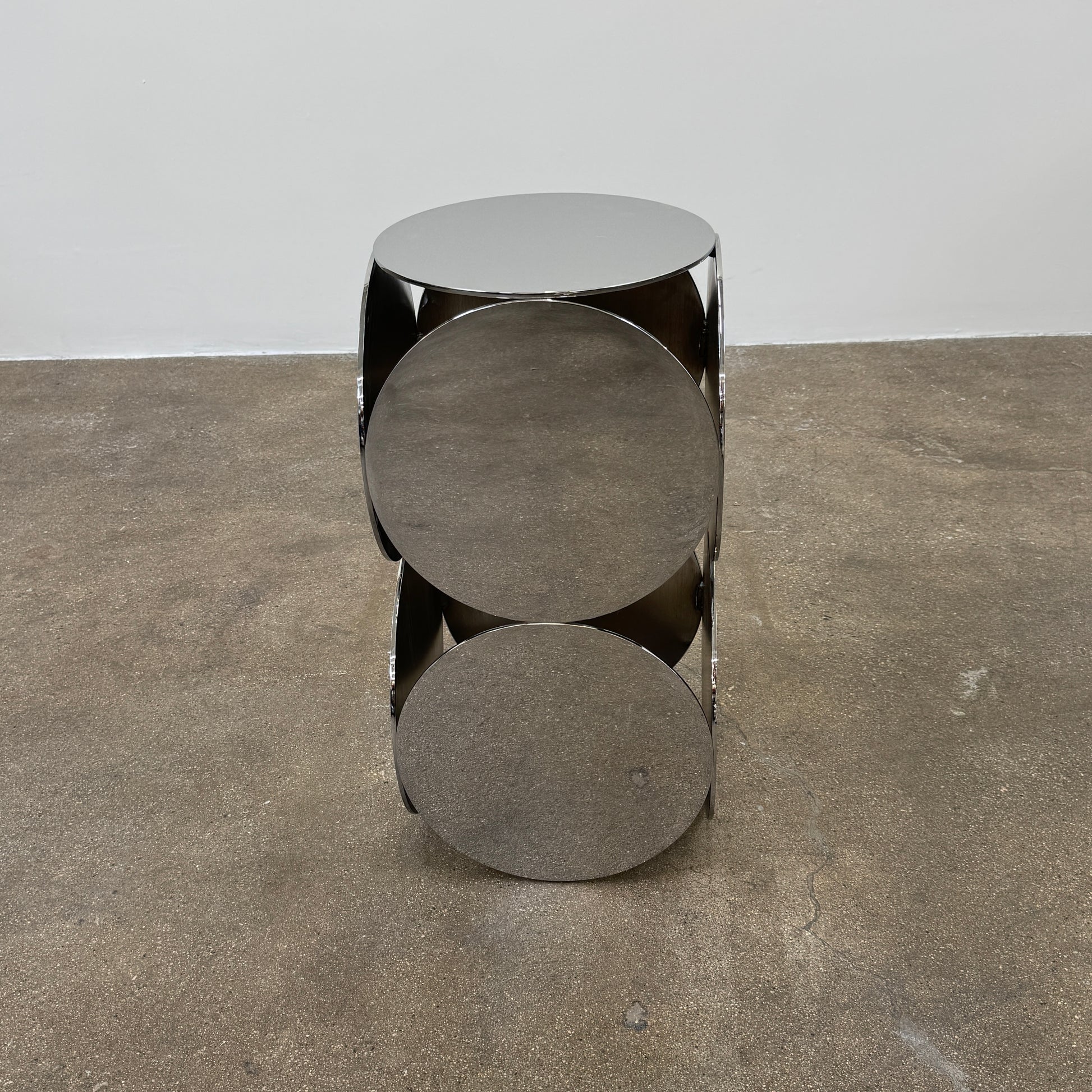 The Minotti Still Accent Table by Minotti features four large, circular, reflective metal discs stacked vertically for minimal elegance, shown on a concrete floor against a white wall.