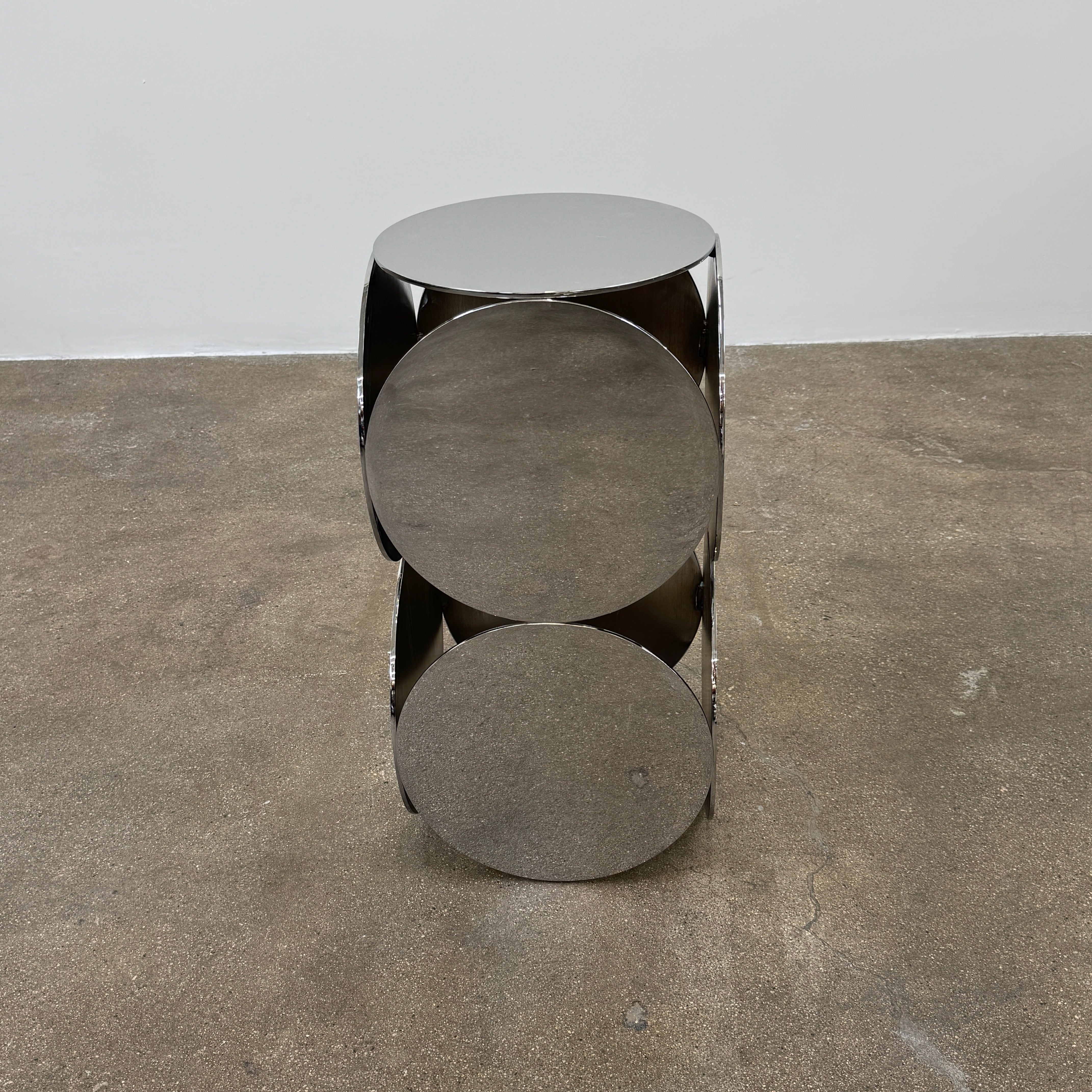 The Minotti Still Accent Table by Minotti features four large, circular, reflective metal discs stacked vertically for minimal elegance, shown on a concrete floor against a white wall.