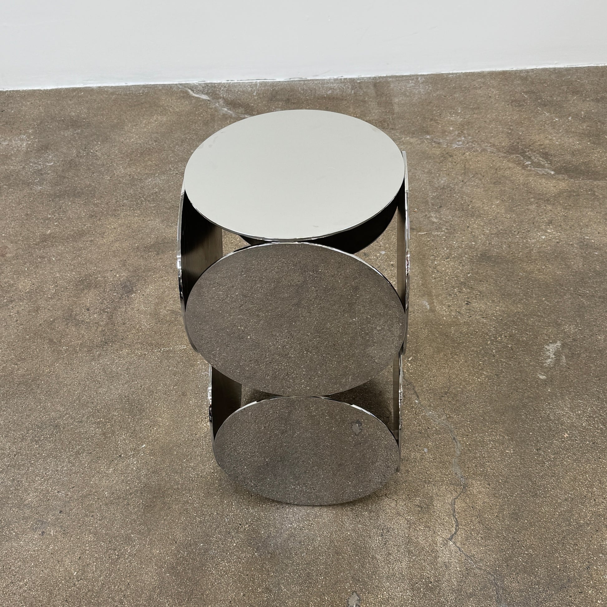 The Minotti Still Accent Table by Minotti features four large, circular, reflective metal discs stacked vertically for minimal elegance, shown on a concrete floor against a white wall.