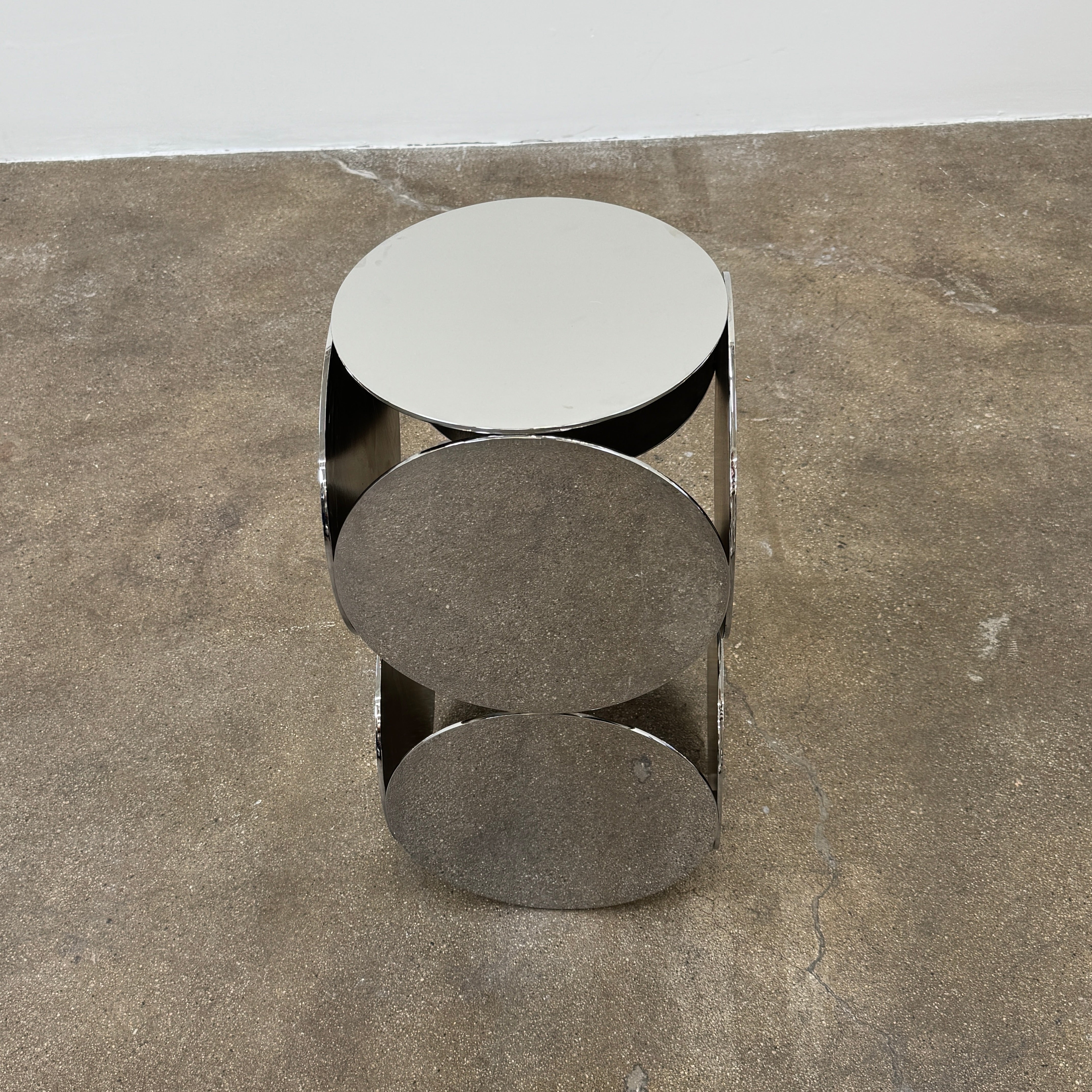 The Minotti Still Accent Table by Minotti features four large, circular, reflective metal discs stacked vertically for minimal elegance, shown on a concrete floor against a white wall.