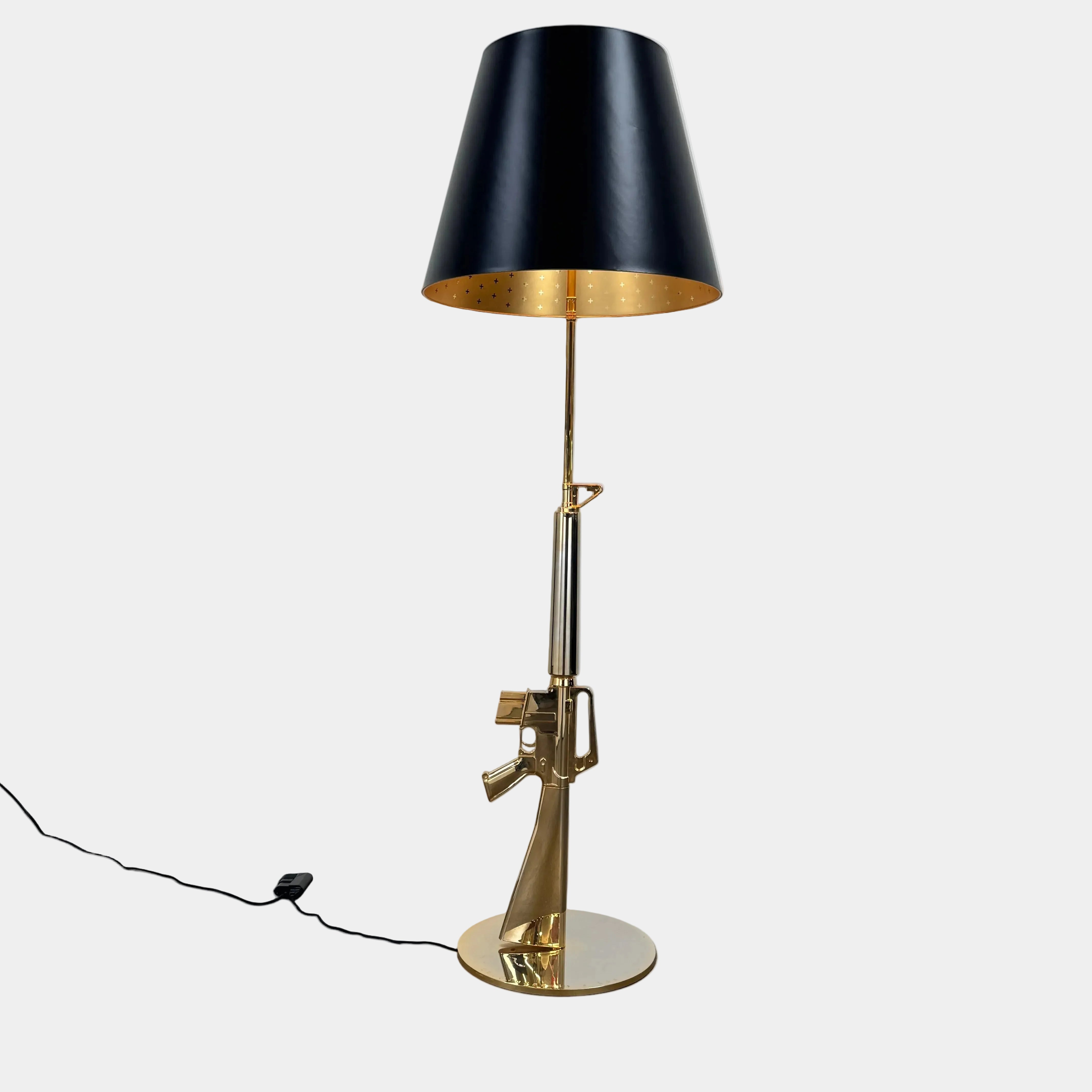 The Flos Guns Lounge Floor Lamp Gold by Flos features a shiny gold rifle-shaped base and a black lampshade, inspired by Philippe Starck, standing against a plain white background.