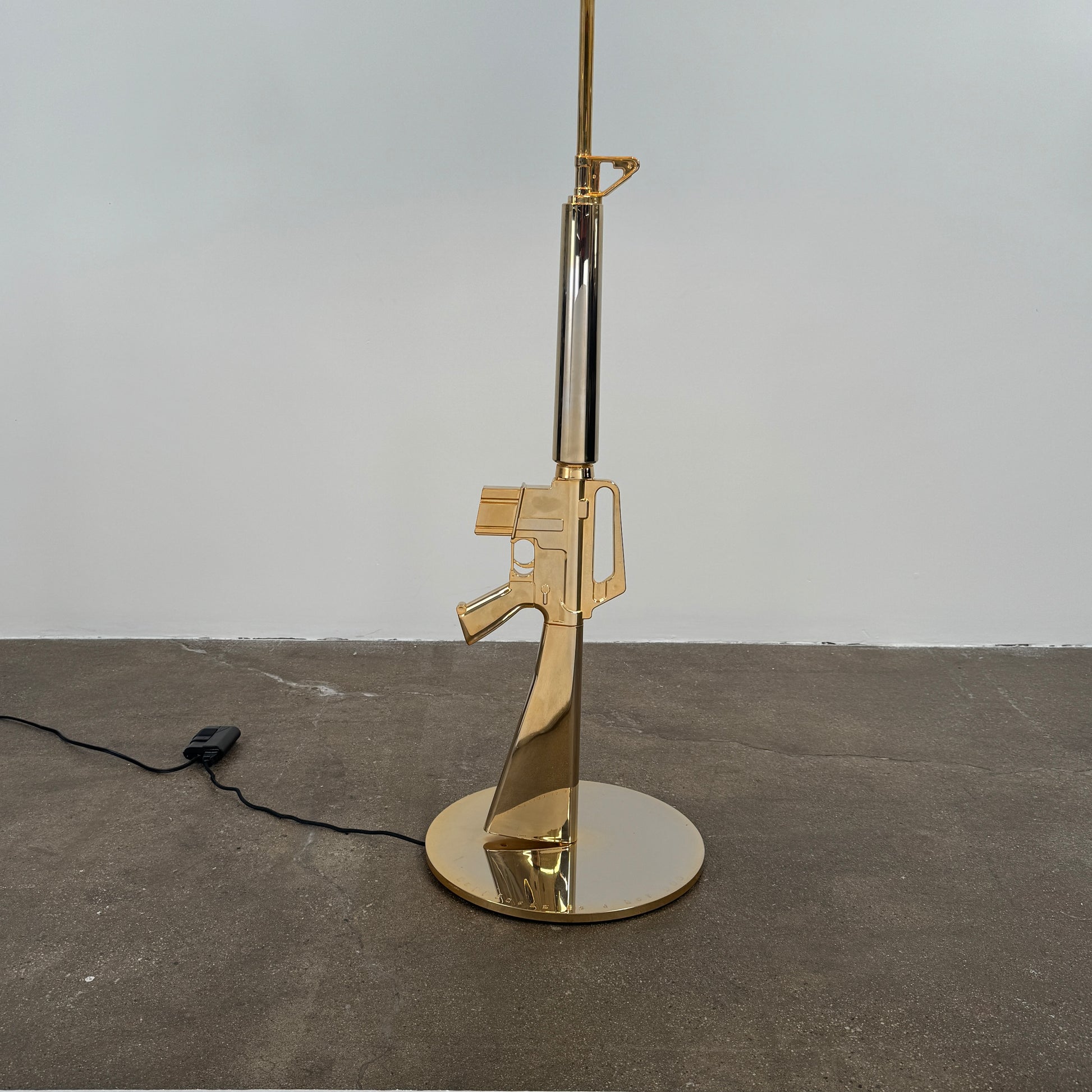 The Flos Guns Lounge Floor Lamp Gold by Flos features a shiny gold rifle-shaped base and a black lampshade, inspired by Philippe Starck, standing against a plain white background.