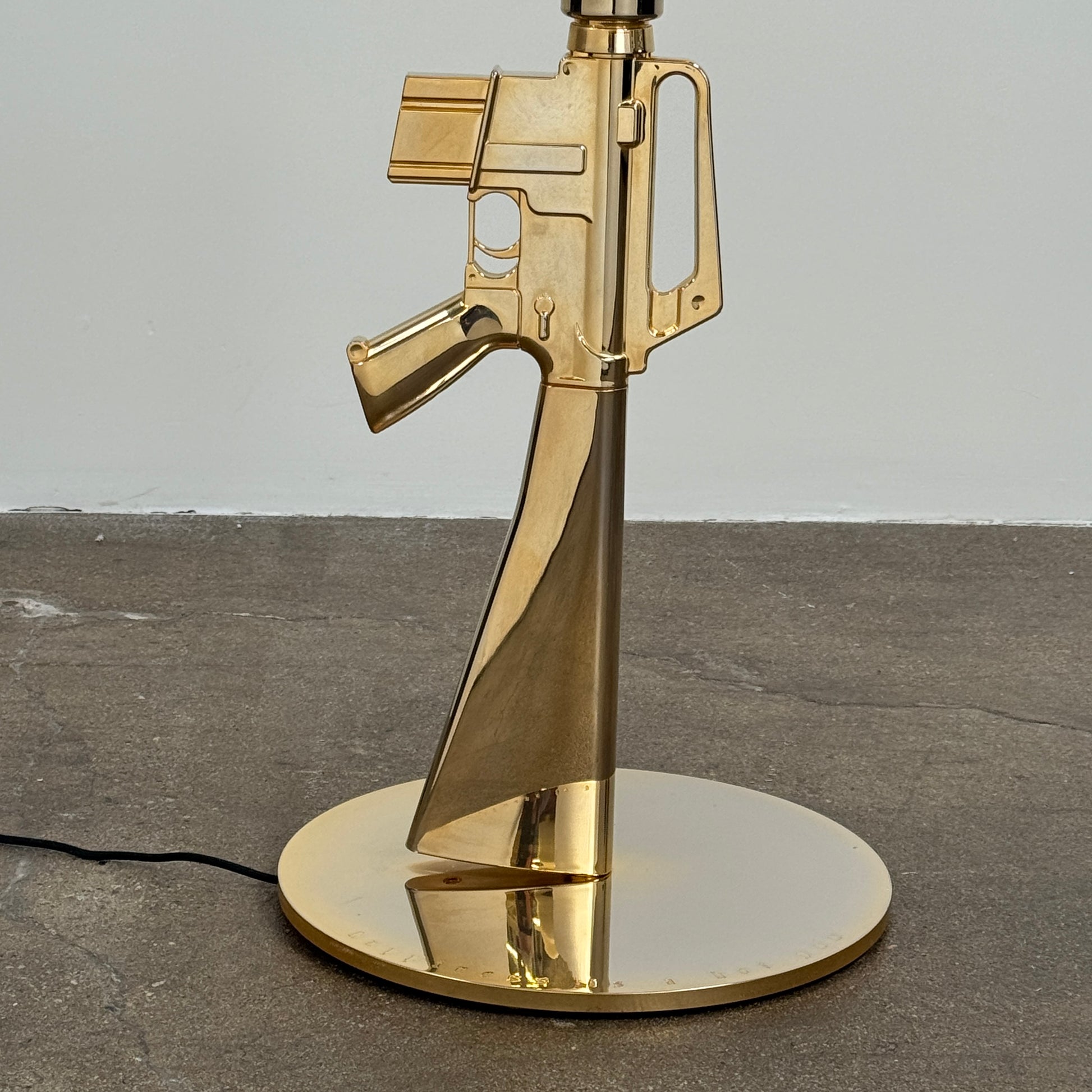 The Flos Guns Lounge Floor Lamp Gold by Flos features a shiny gold rifle-shaped base and a black lampshade, inspired by Philippe Starck, standing against a plain white background.