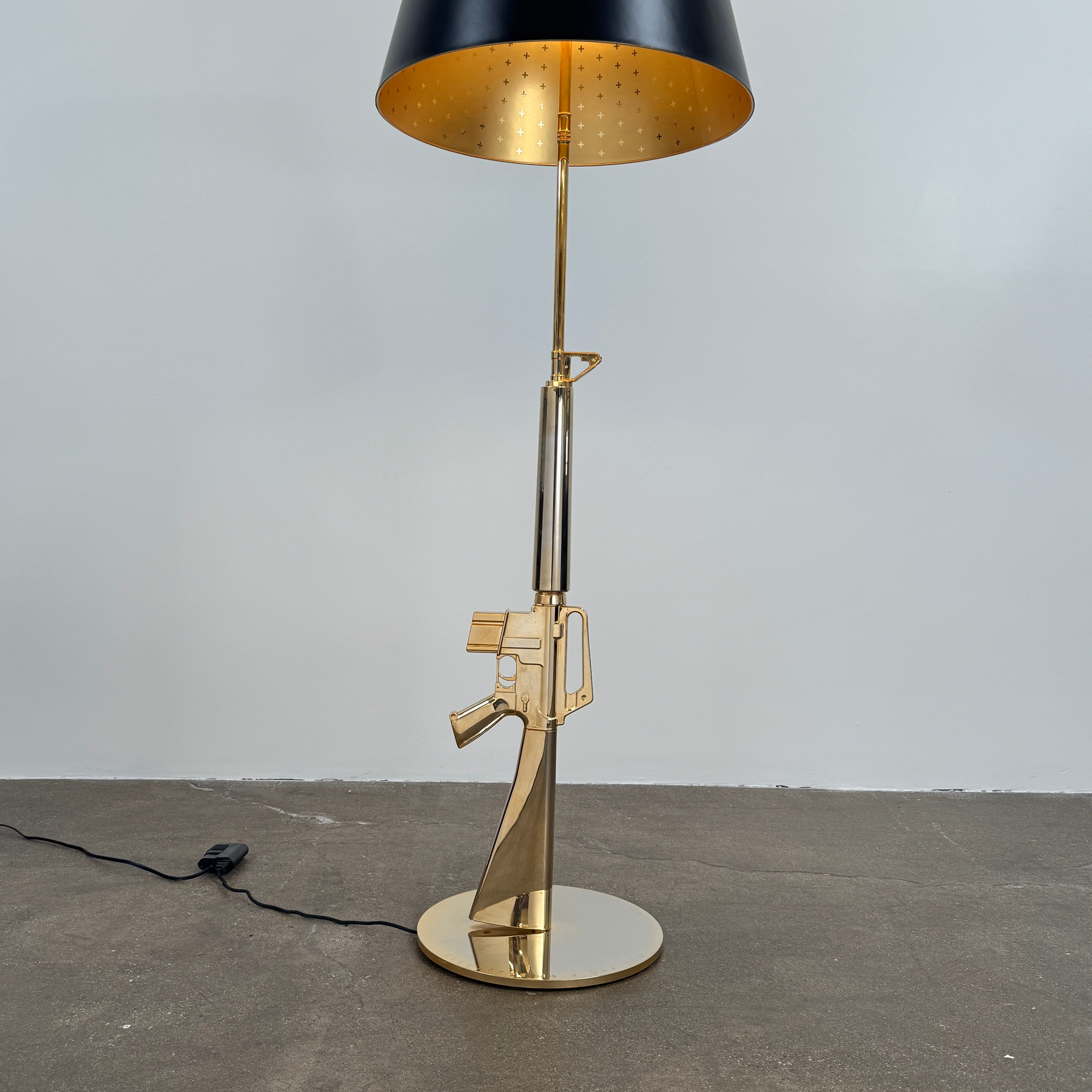 The Flos Guns Lounge Floor Lamp Gold by Flos features a shiny gold rifle-shaped base and a black lampshade, inspired by Philippe Starck, standing against a plain white background.