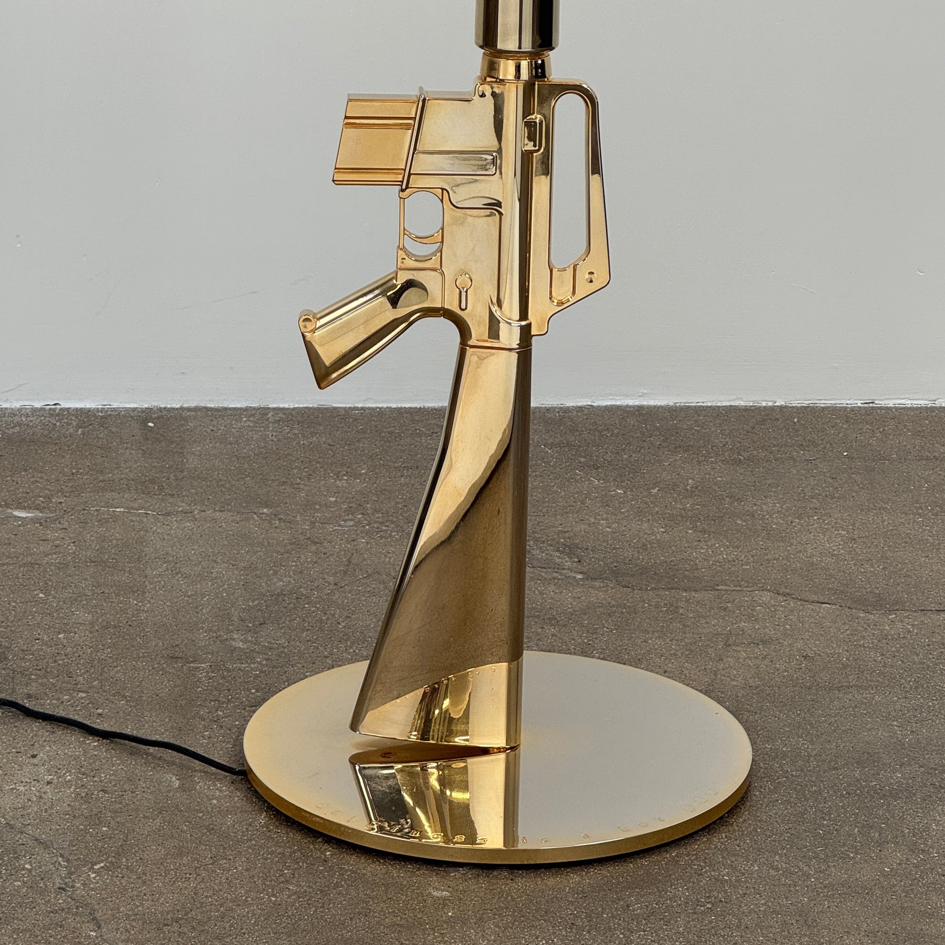 The Flos Guns Lounge Floor Lamp Gold by Flos features a shiny gold rifle-shaped base and a black lampshade, inspired by Philippe Starck, standing against a plain white background.