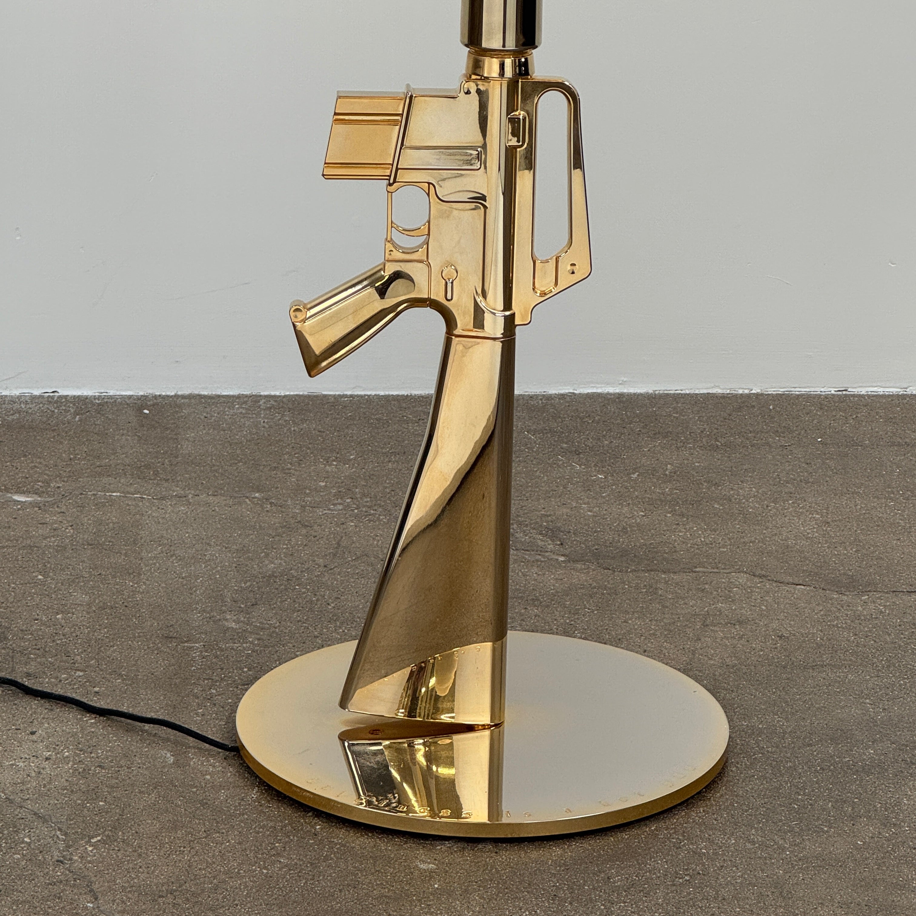 The Flos Guns Lounge Floor Lamp Gold by Flos features a shiny gold rifle-shaped base and a black lampshade, inspired by Philippe Starck, standing against a plain white background.