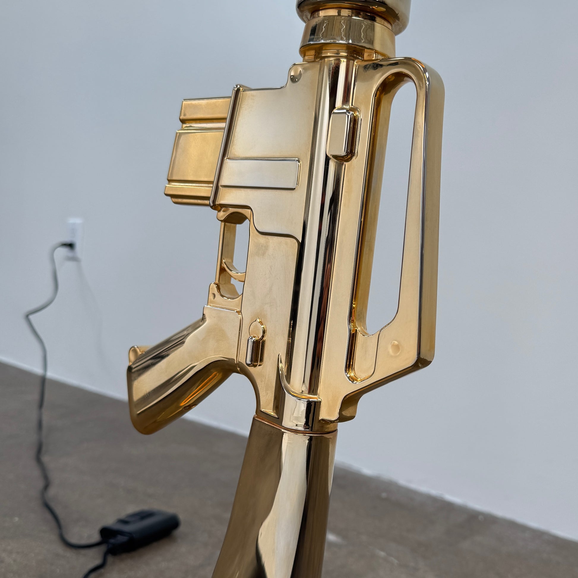 The Flos Guns Lounge Floor Lamp Gold by Flos features a shiny gold rifle-shaped base and a black lampshade, inspired by Philippe Starck, standing against a plain white background.