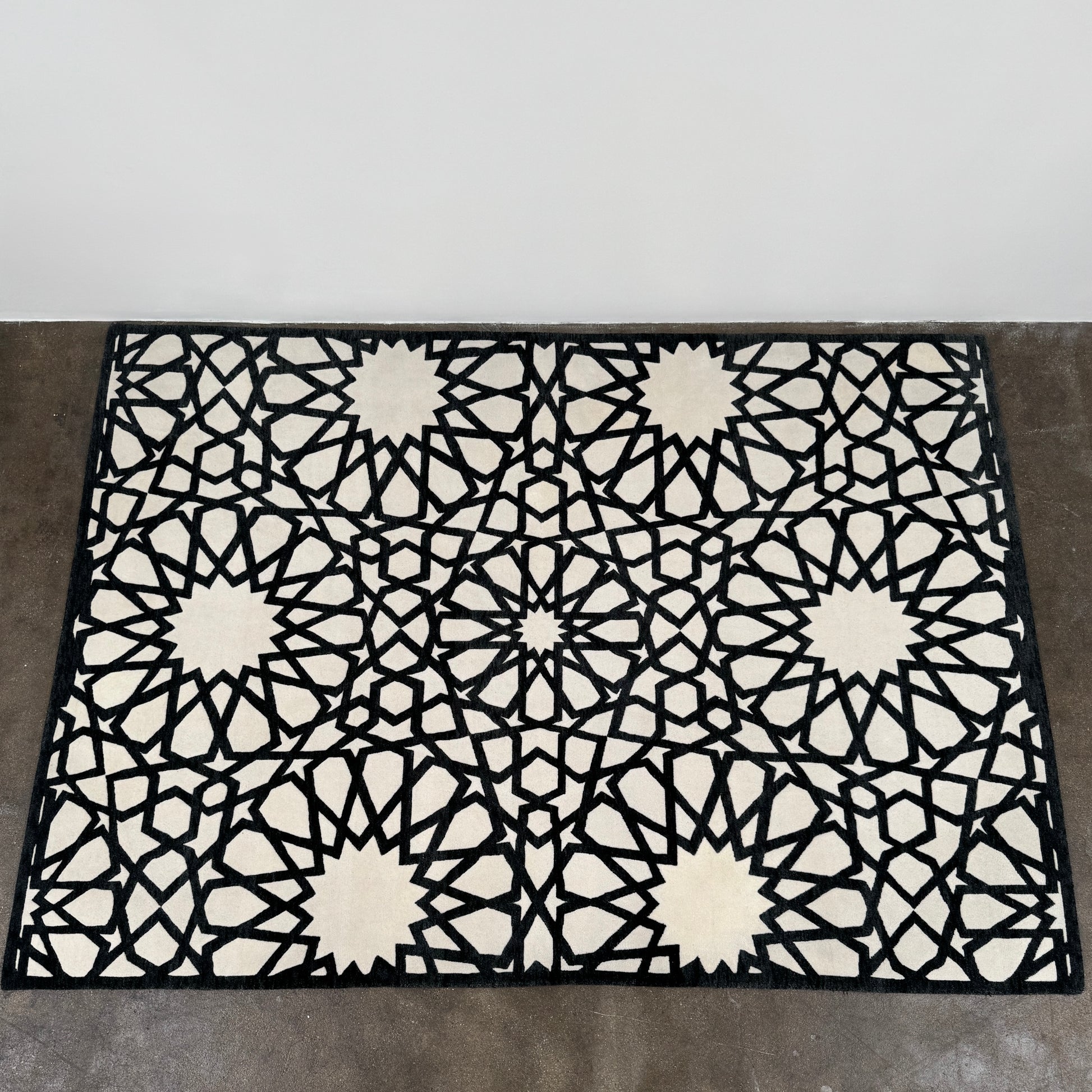 The Roche Bobois Moucharabieh Rug by Roche Bobois features a geometric black and white pattern with interlocking shapes and starbursts, displayed on a plain floor against a white wall.