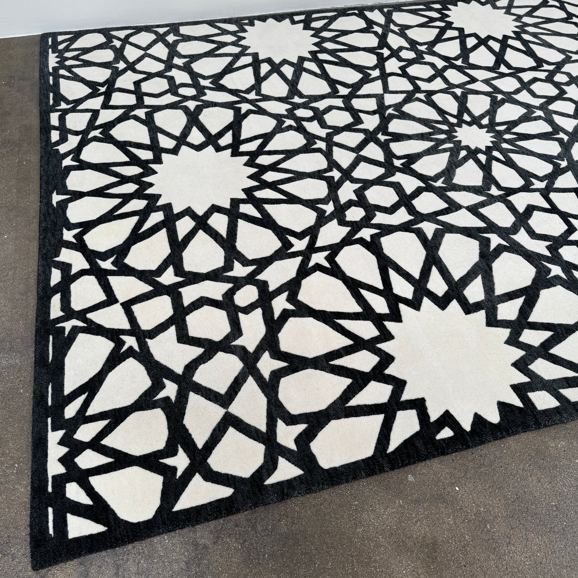 The Roche Bobois Moucharabieh Rug by Roche Bobois features a geometric black and white pattern with interlocking shapes and starbursts, displayed on a plain floor against a white wall.
