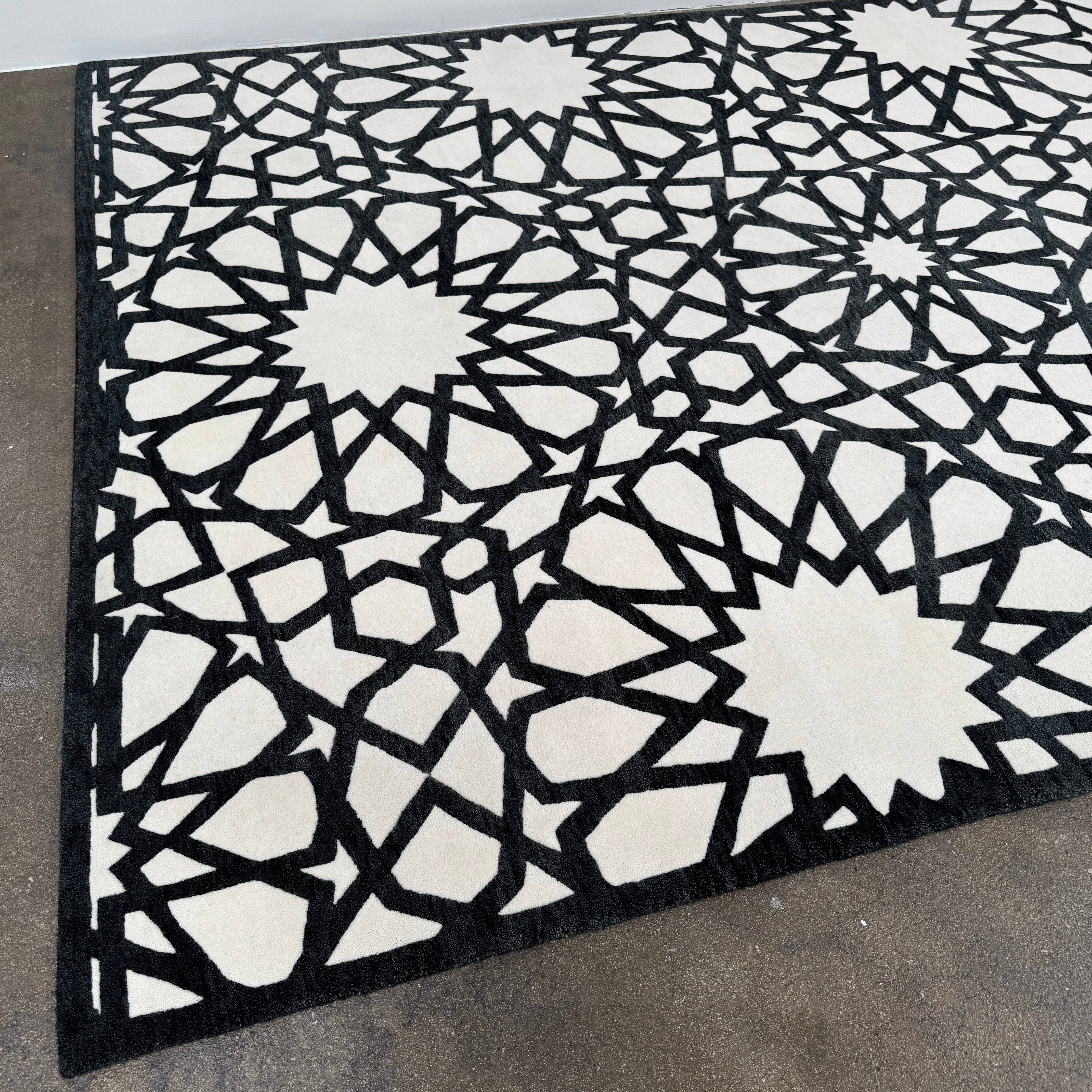The Roche Bobois Moucharabieh Rug by Roche Bobois features a geometric black and white pattern with interlocking shapes and starbursts, displayed on a plain floor against a white wall.
