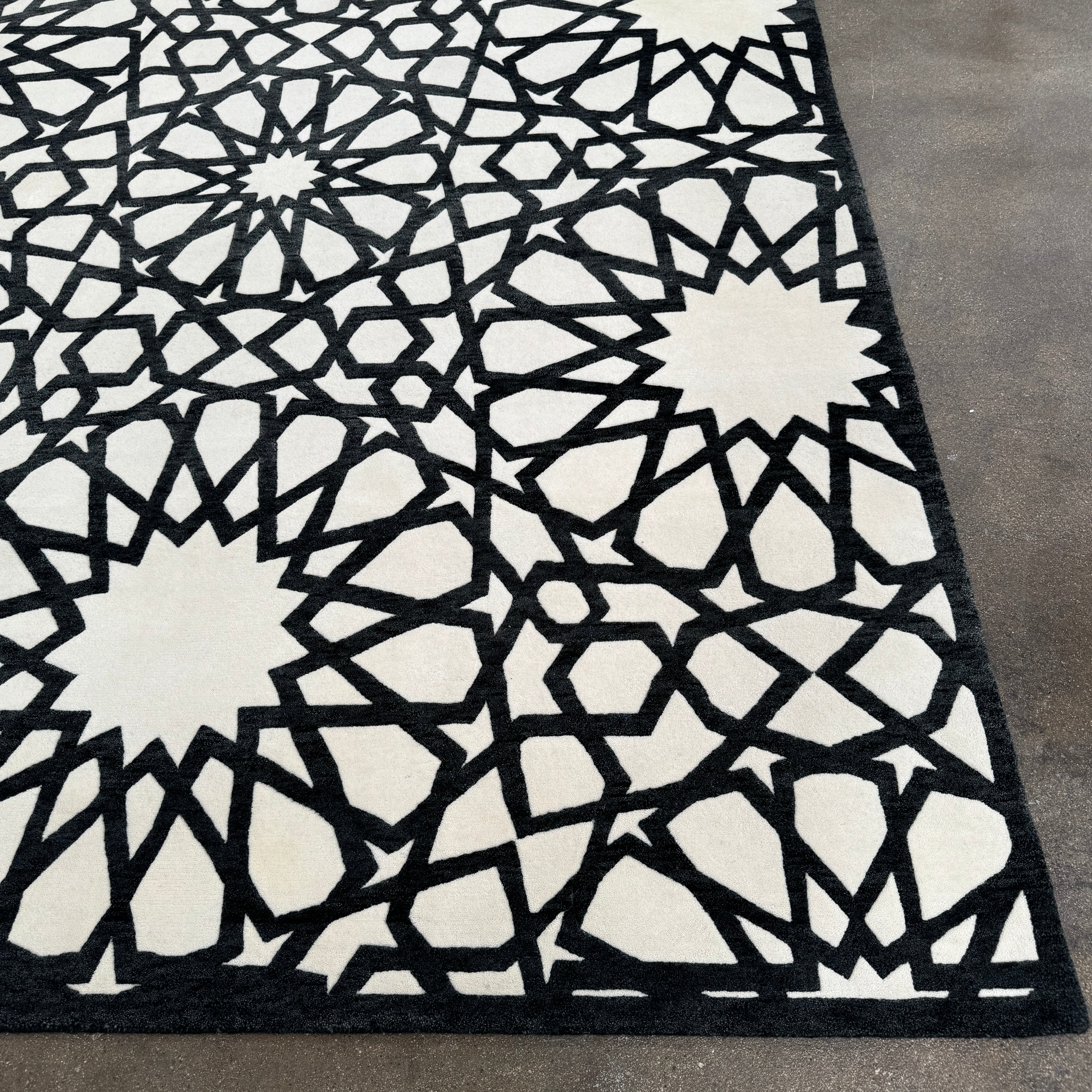 The Roche Bobois Moucharabieh Rug by Roche Bobois features a geometric black and white pattern with interlocking shapes and starbursts, displayed on a plain floor against a white wall.