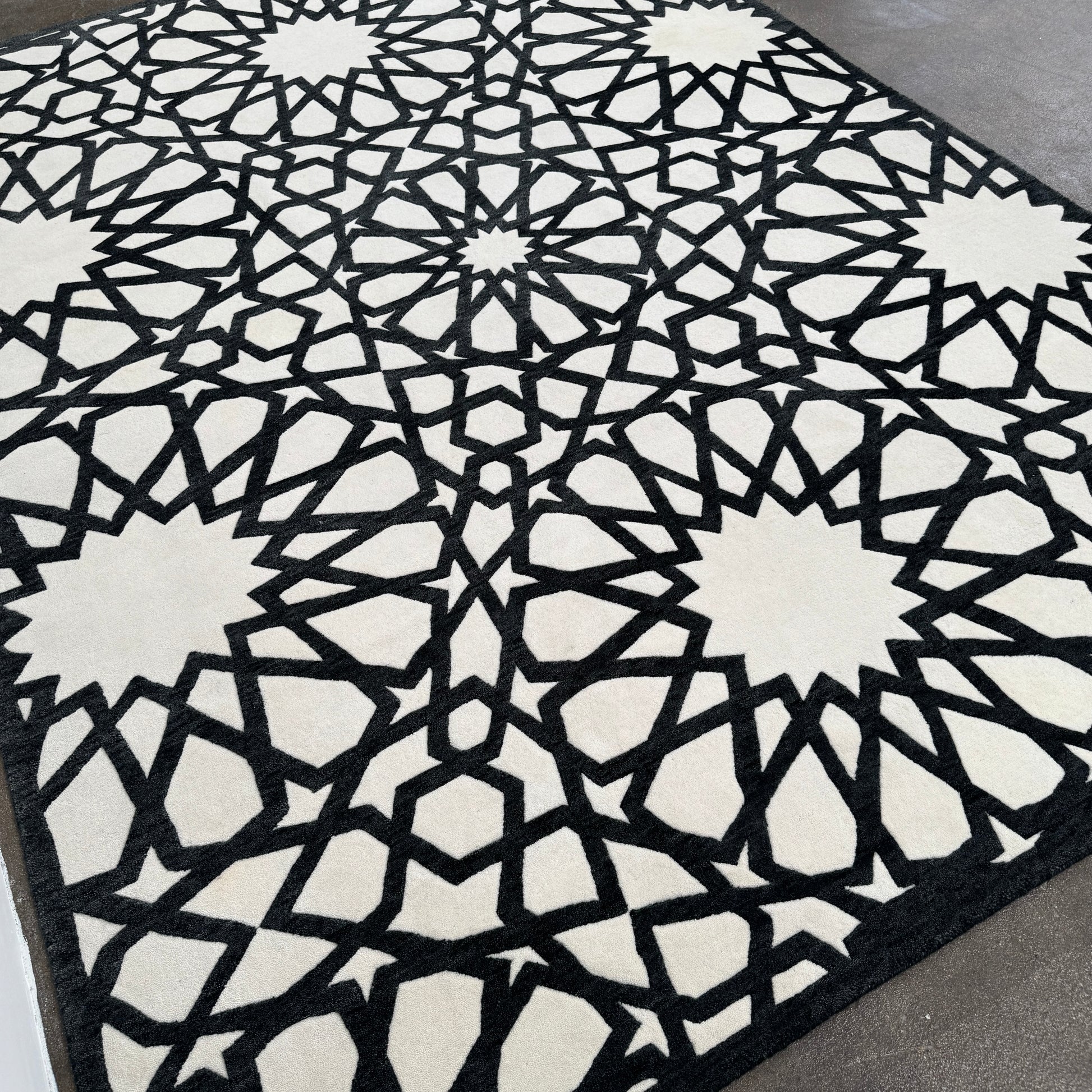 The Roche Bobois Moucharabieh Rug by Roche Bobois features a geometric black and white pattern with interlocking shapes and starbursts, displayed on a plain floor against a white wall.