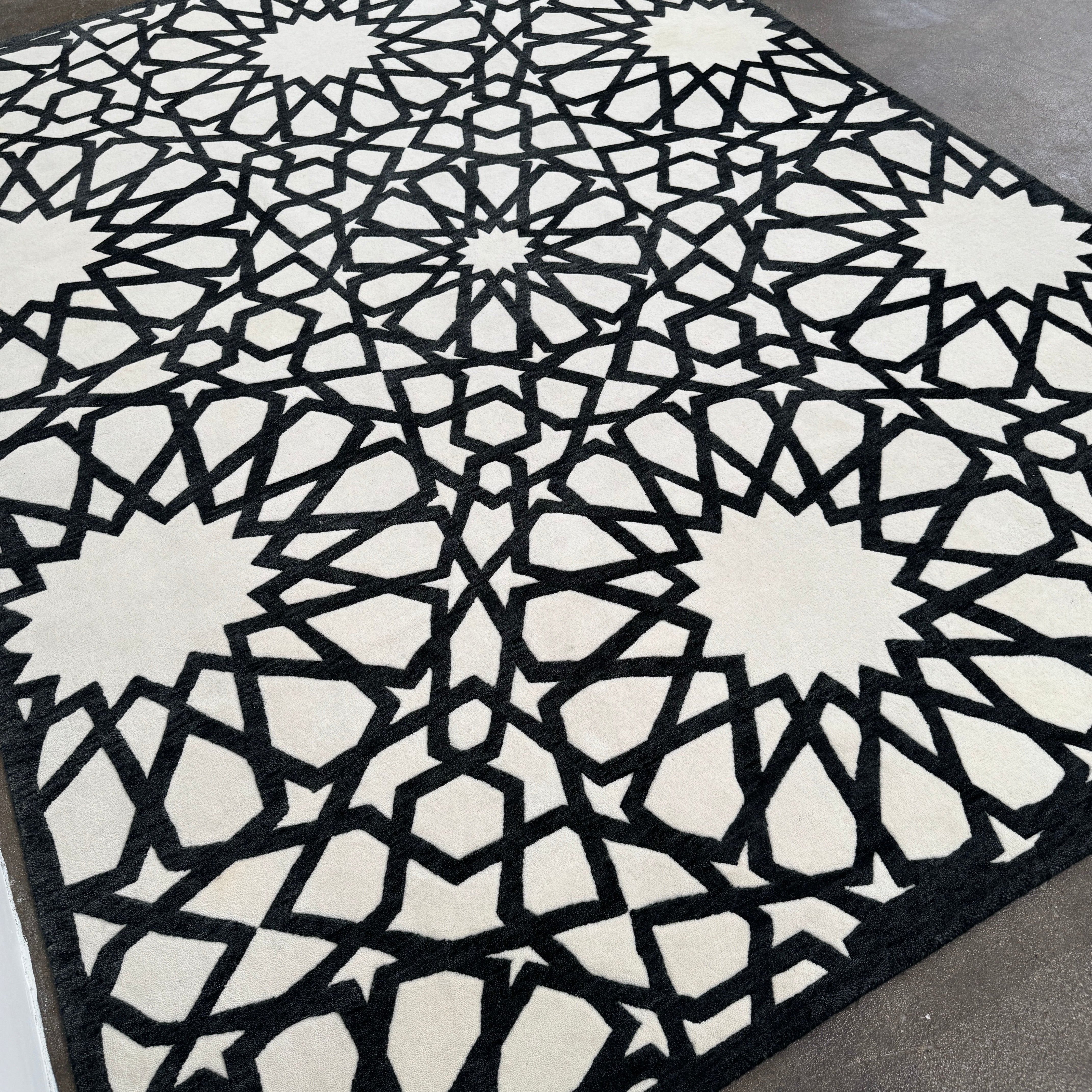 The Roche Bobois Moucharabieh Rug by Roche Bobois features a geometric black and white pattern with interlocking shapes and starbursts, displayed on a plain floor against a white wall.
