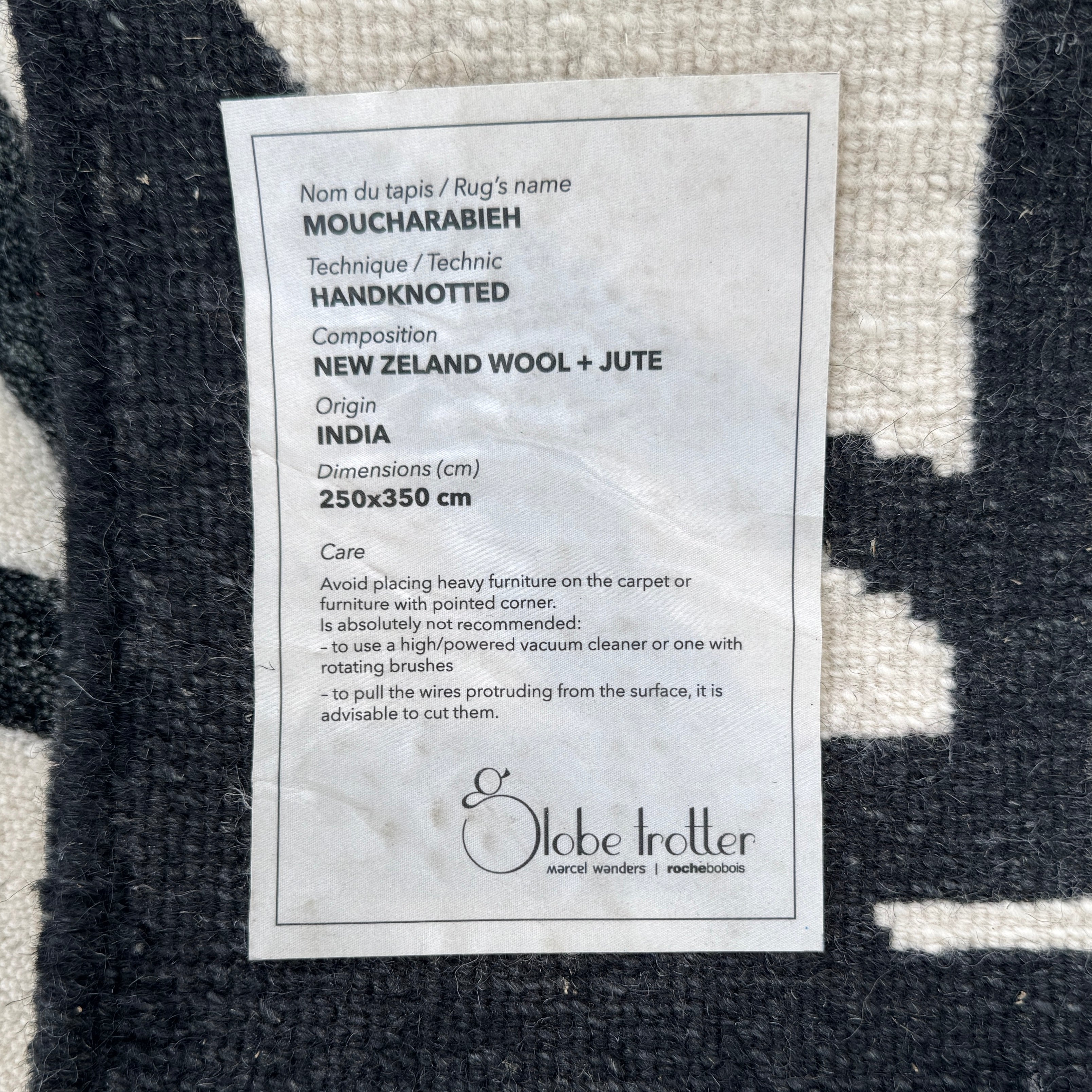 The Roche Bobois Moucharabieh Rug by Roche Bobois features a geometric black and white pattern with interlocking shapes and starbursts, displayed on a plain floor against a white wall.