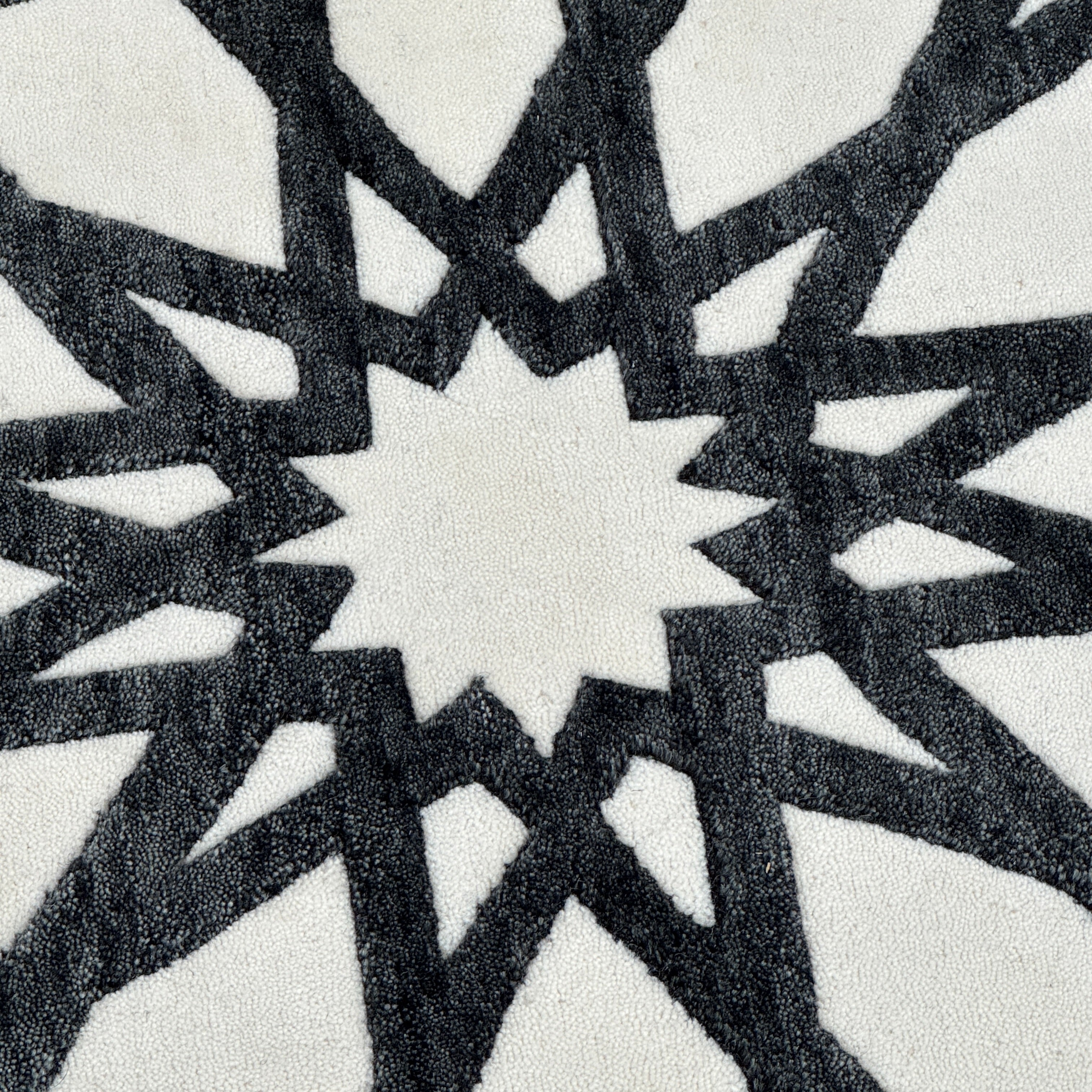 The Roche Bobois Moucharabieh Rug by Roche Bobois features a geometric black and white pattern with interlocking shapes and starbursts, displayed on a plain floor against a white wall.