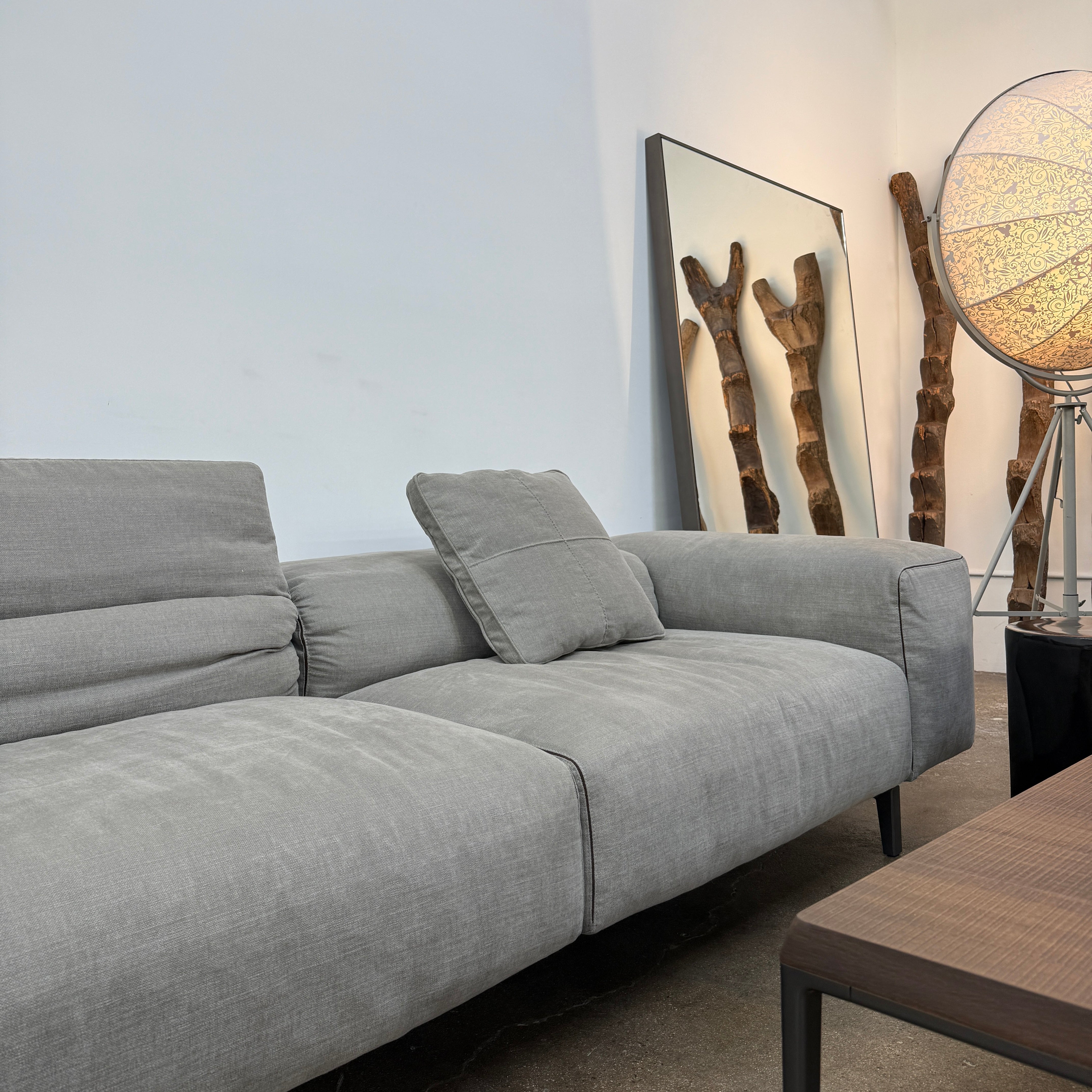 A modern Cassina Scighera Sofa by Cassina, in light gray with two matching square cushions, is placed on a bare concrete floor against a plain white wall.