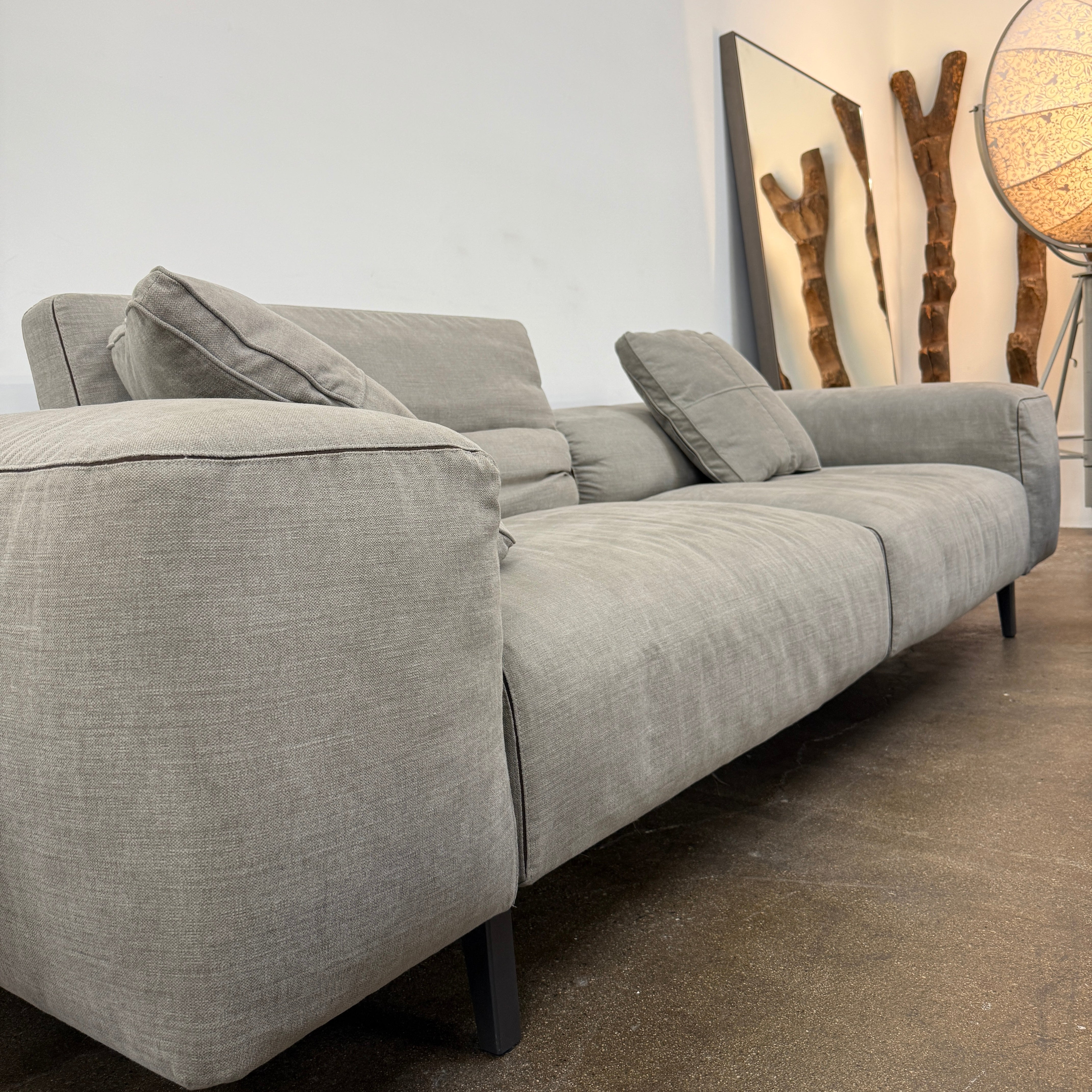 A modern Cassina Scighera Sofa by Cassina, in light gray with two matching square cushions, is placed on a bare concrete floor against a plain white wall.