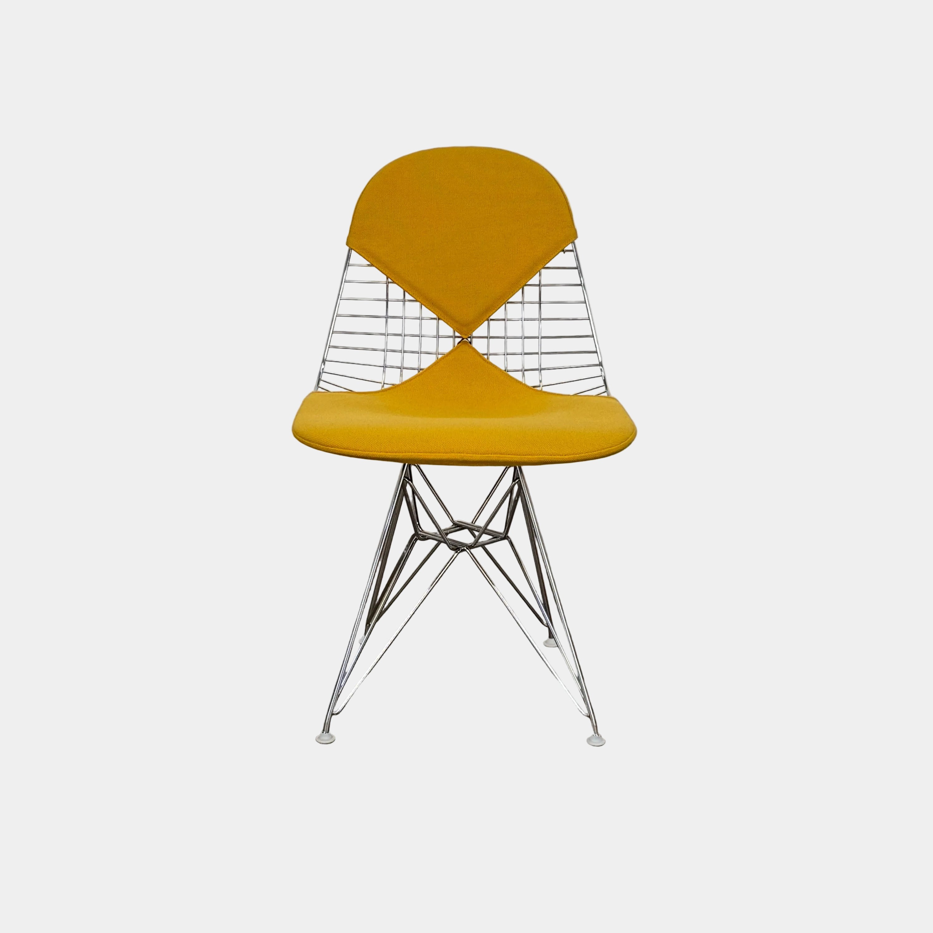 The Herman Miller Eames Wire Chair with Bikini Pad by Herman Miller features a yellow cushioned seat and backrest, metal wire frame with Eiffel Tower base, and angled legs, set on a concrete floor against a plain white wall.