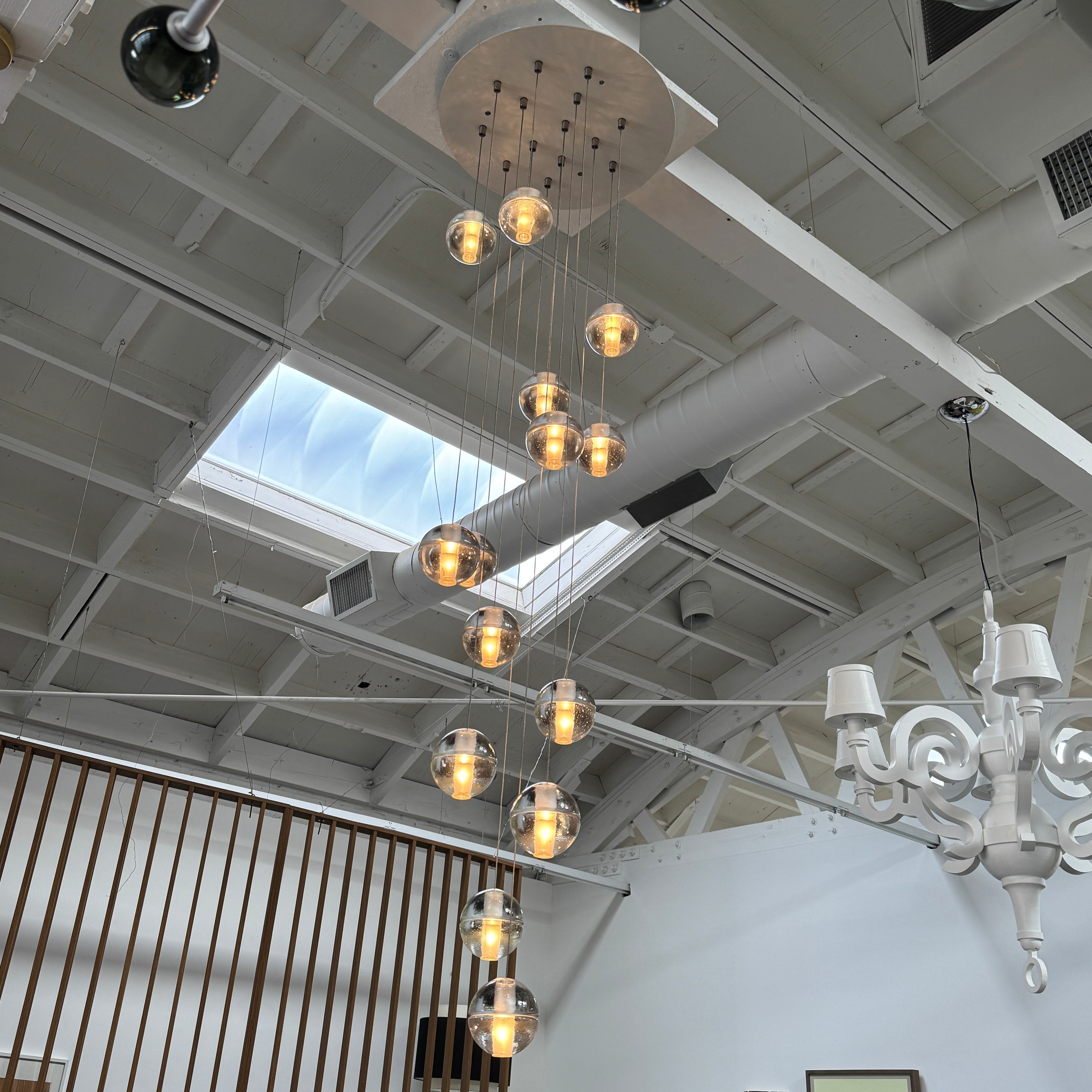 The Bocci 14.14 Pendant Chandelier by Bocci features a vertical cluster of metallic pendants, each with a glowing bulb, suspended by thin cords against a white background for a modern look.