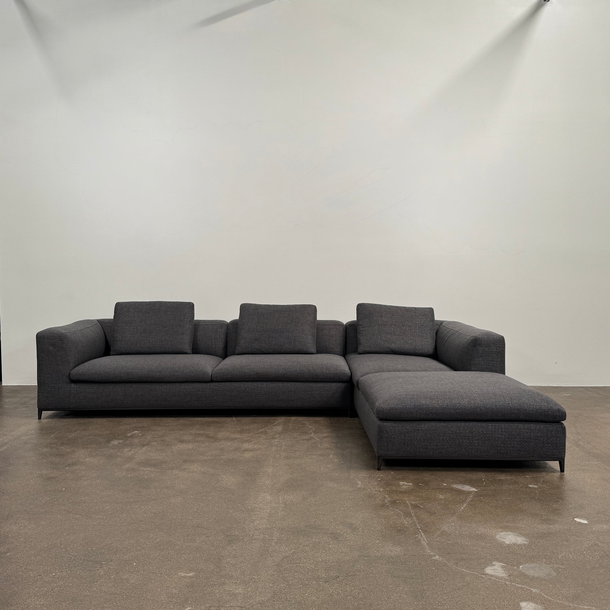 The B&B Italia Michele Club Sectional Sofa by B&B Italia, in dark grey open weave fabric with three seats and a right-side chaise lounge, sits on a concrete floor against a white wall—perfect for modern living spaces.