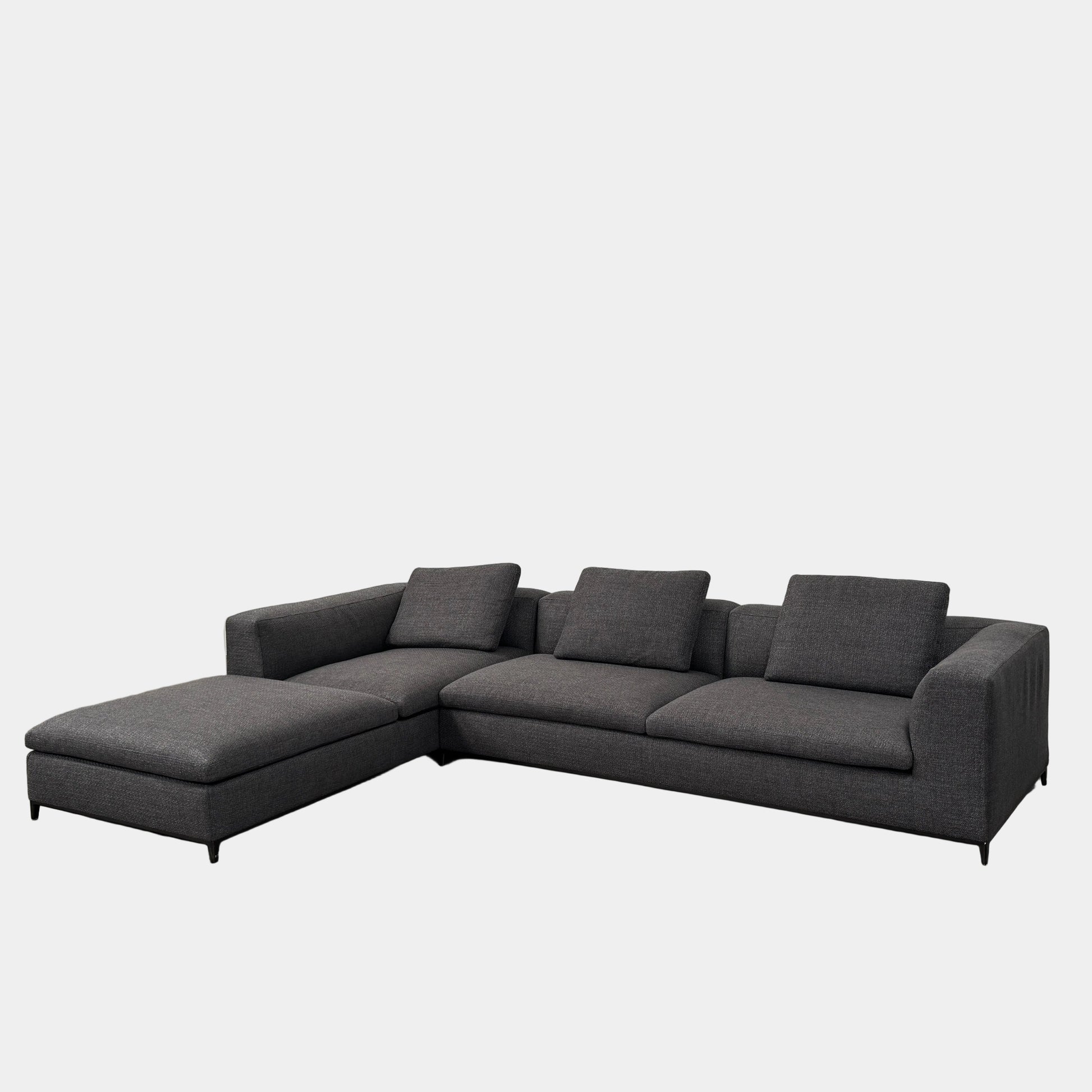 The B&B Italia Michele Club Sectional Sofa by B&B Italia, in dark grey open weave fabric with three seats and a right-side chaise lounge, sits on a concrete floor against a white wall—perfect for modern living spaces.