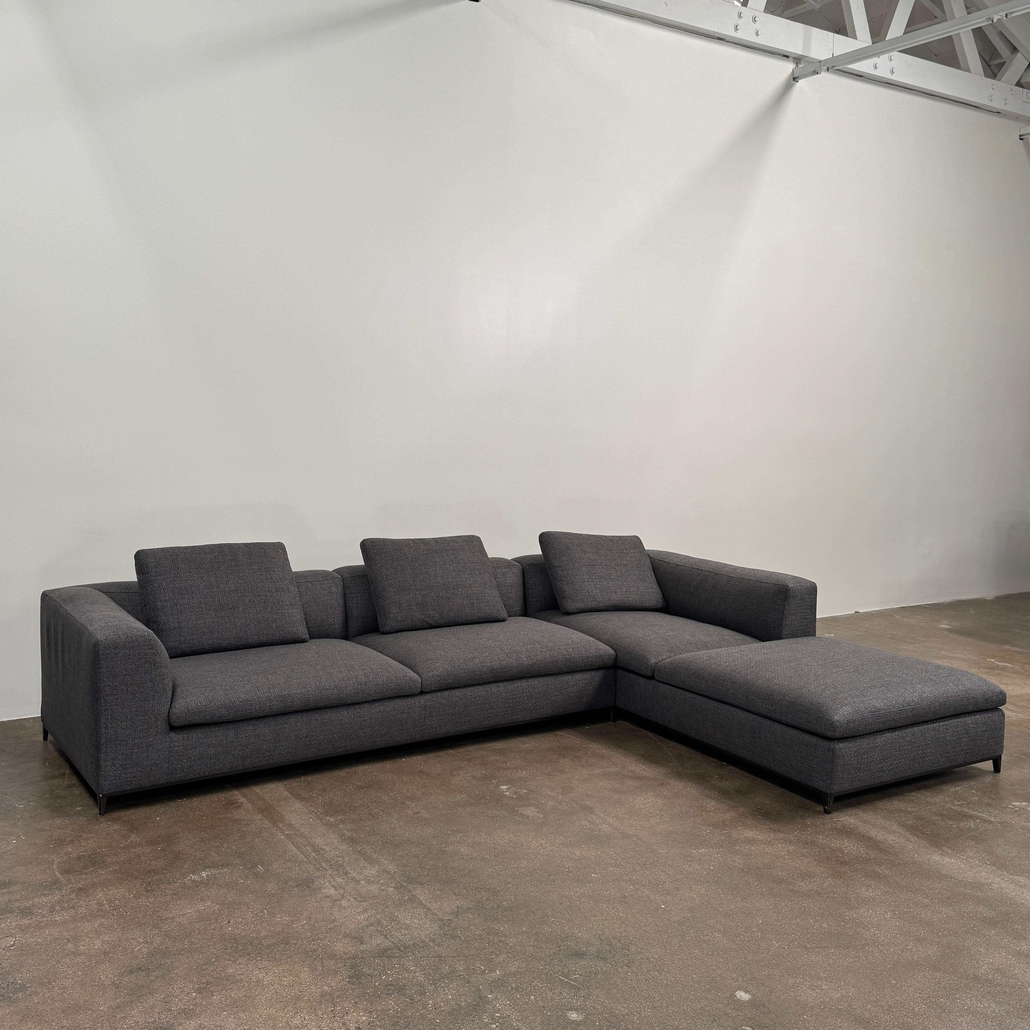 The B&B Italia Michele Club Sectional Sofa by B&B Italia, in dark grey open weave fabric with three seats and a right-side chaise lounge, sits on a concrete floor against a white wall—perfect for modern living spaces.