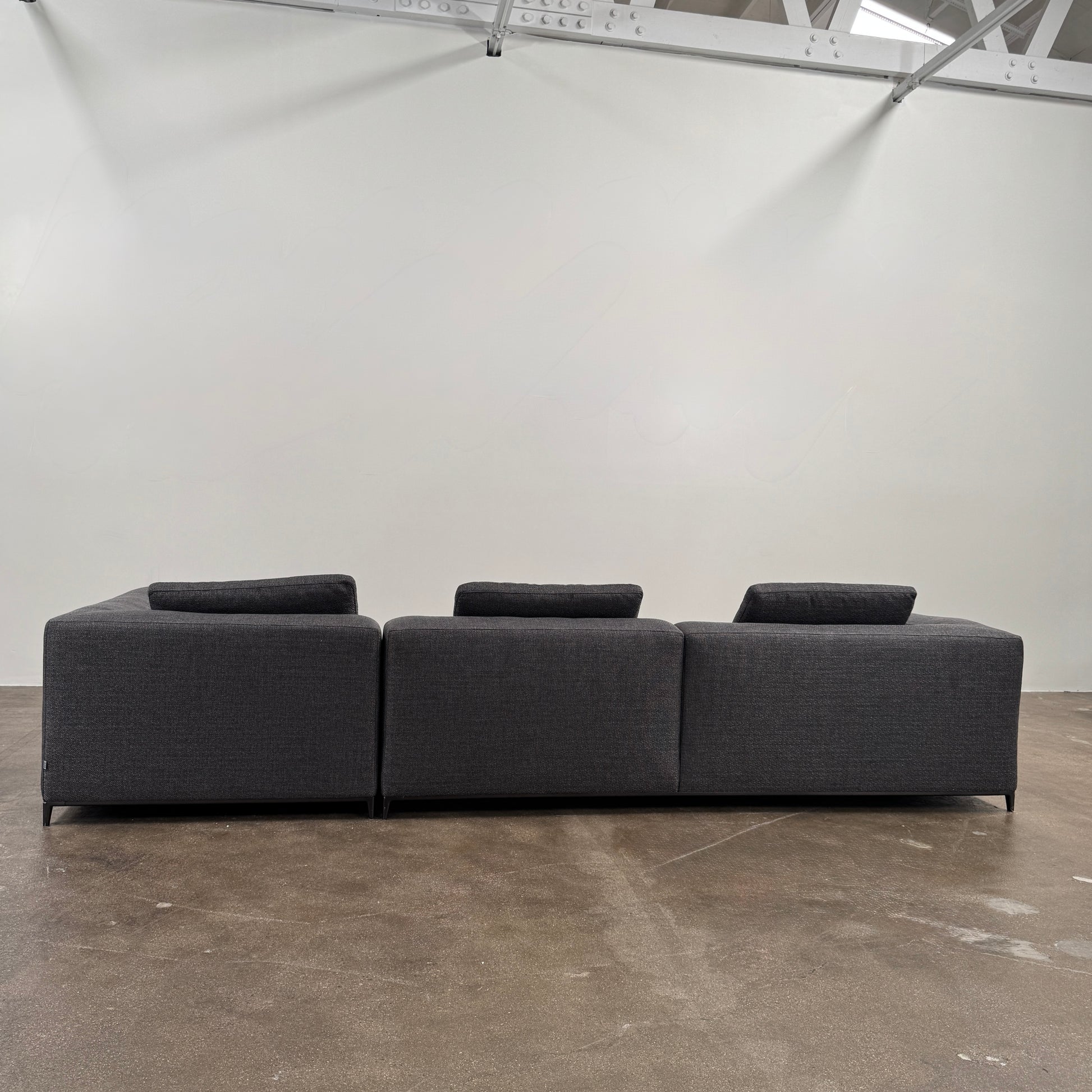 The B&B Italia Michele Club Sectional Sofa by B&B Italia, in dark grey open weave fabric with three seats and a right-side chaise lounge, sits on a concrete floor against a white wall—perfect for modern living spaces.
