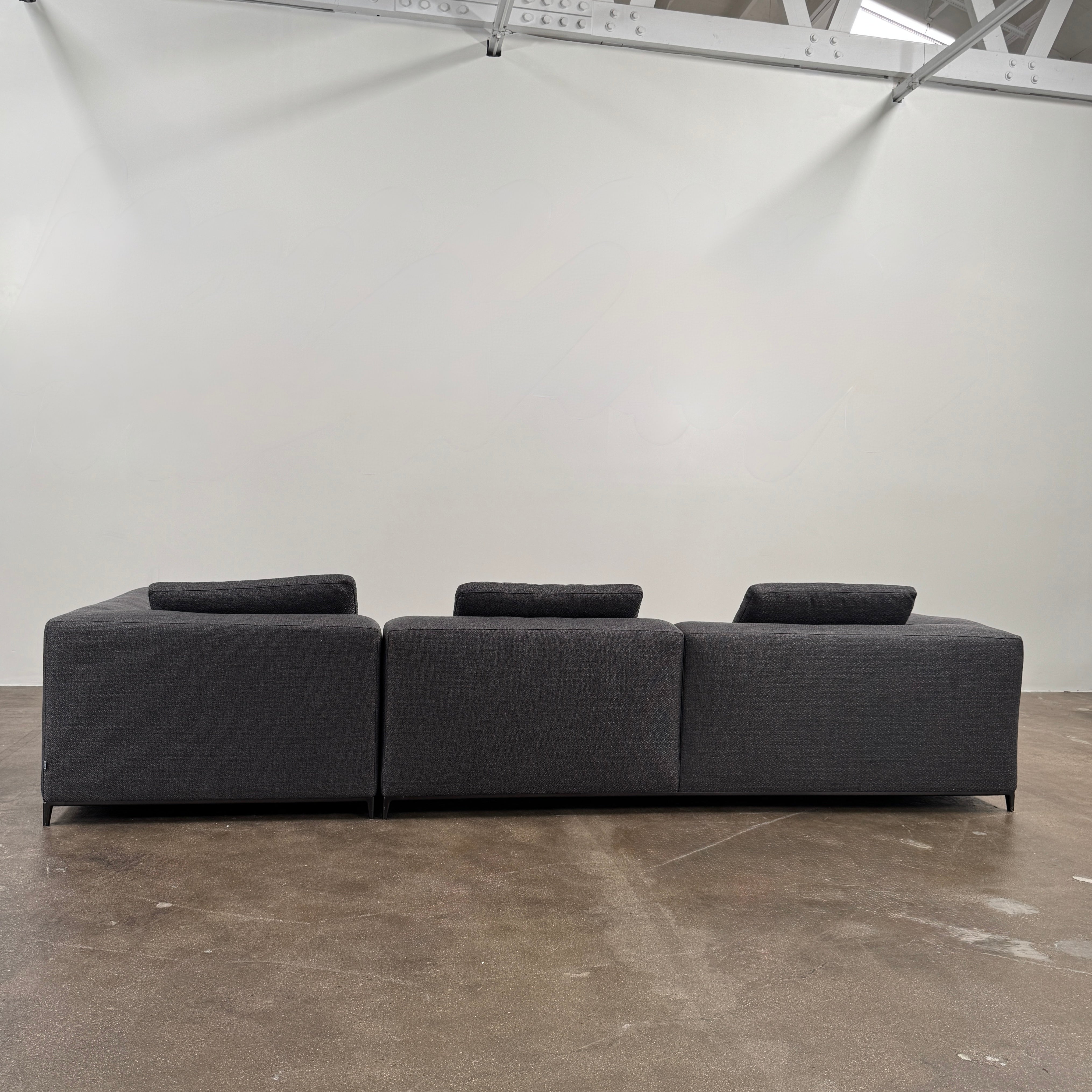 The B&B Italia Michele Club Sectional Sofa by B&B Italia, in dark grey open weave fabric with three seats and a right-side chaise lounge, sits on a concrete floor against a white wall—perfect for modern living spaces.