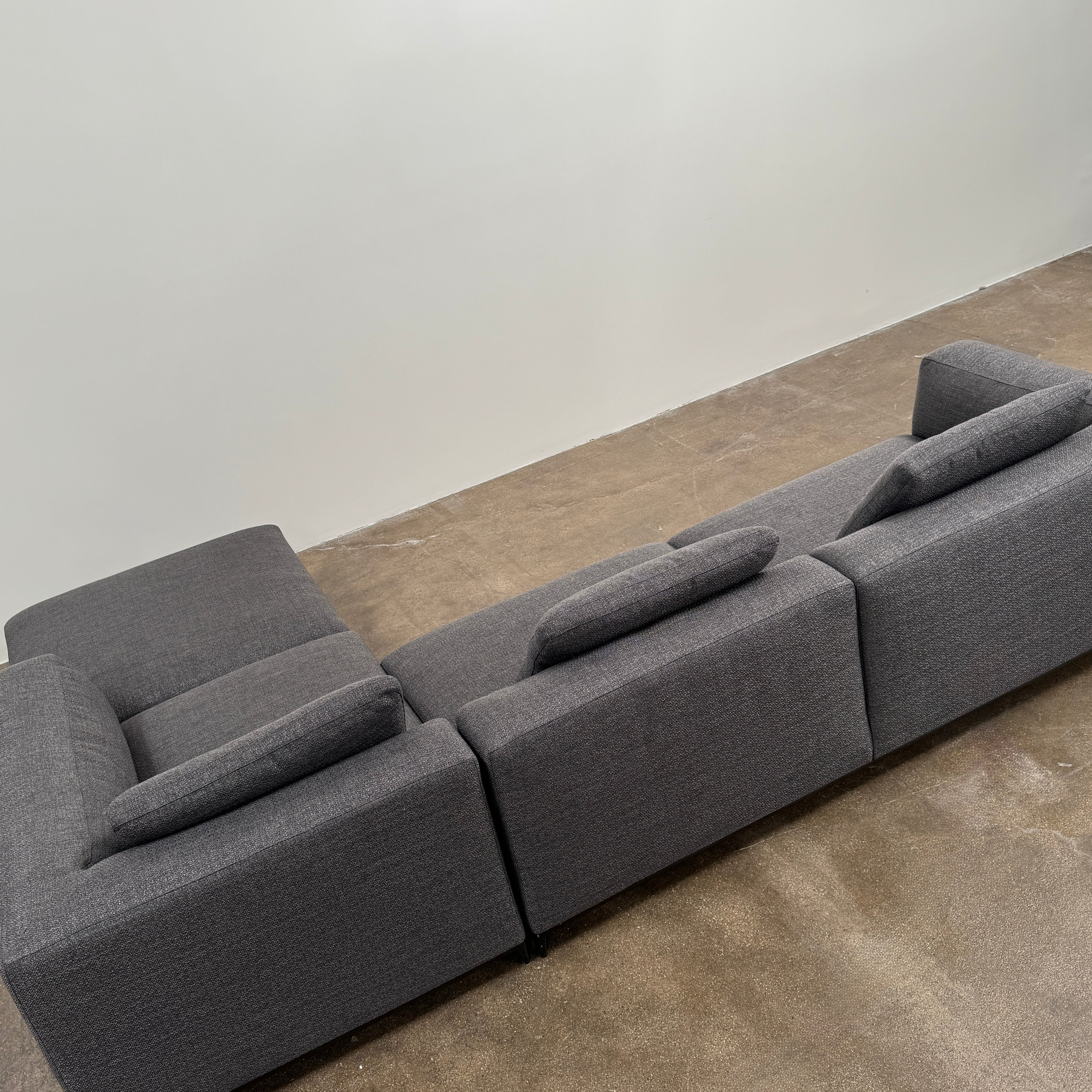 The B&B Italia Michele Club Sectional Sofa by B&B Italia, in dark grey open weave fabric with three seats and a right-side chaise lounge, sits on a concrete floor against a white wall—perfect for modern living spaces.