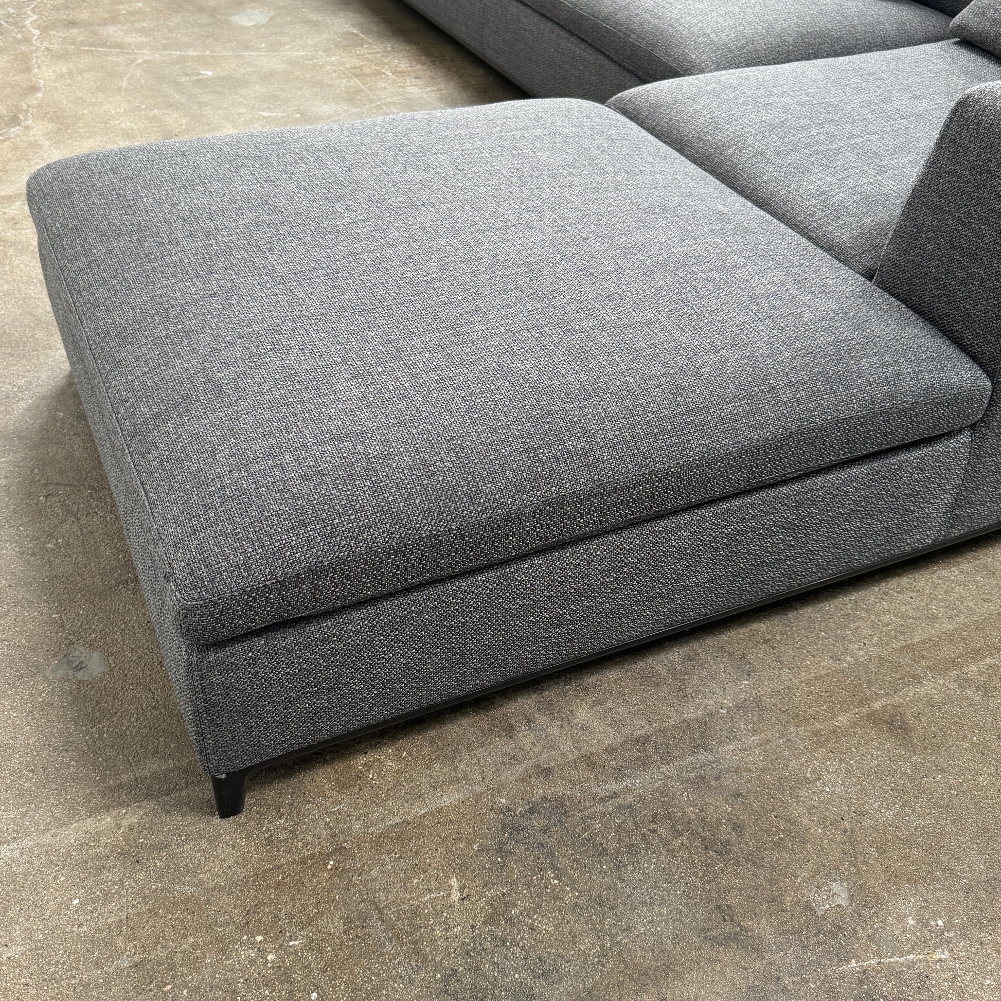 The B&B Italia Michele Club Sectional Sofa by B&B Italia, in dark grey open weave fabric with three seats and a right-side chaise lounge, sits on a concrete floor against a white wall—perfect for modern living spaces.