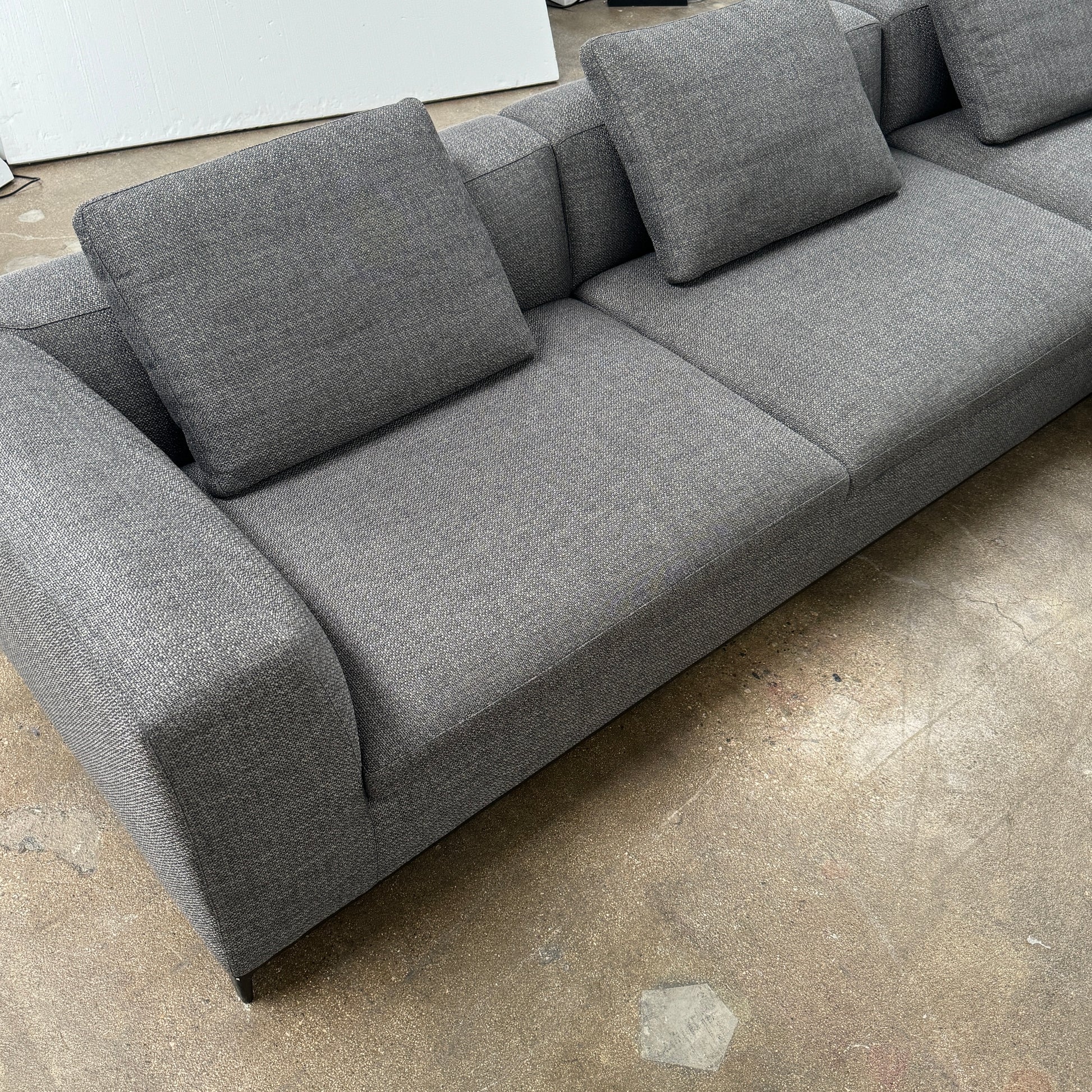 The B&B Italia Michele Club Sectional Sofa by B&B Italia, in dark grey open weave fabric with three seats and a right-side chaise lounge, sits on a concrete floor against a white wall—perfect for modern living spaces.