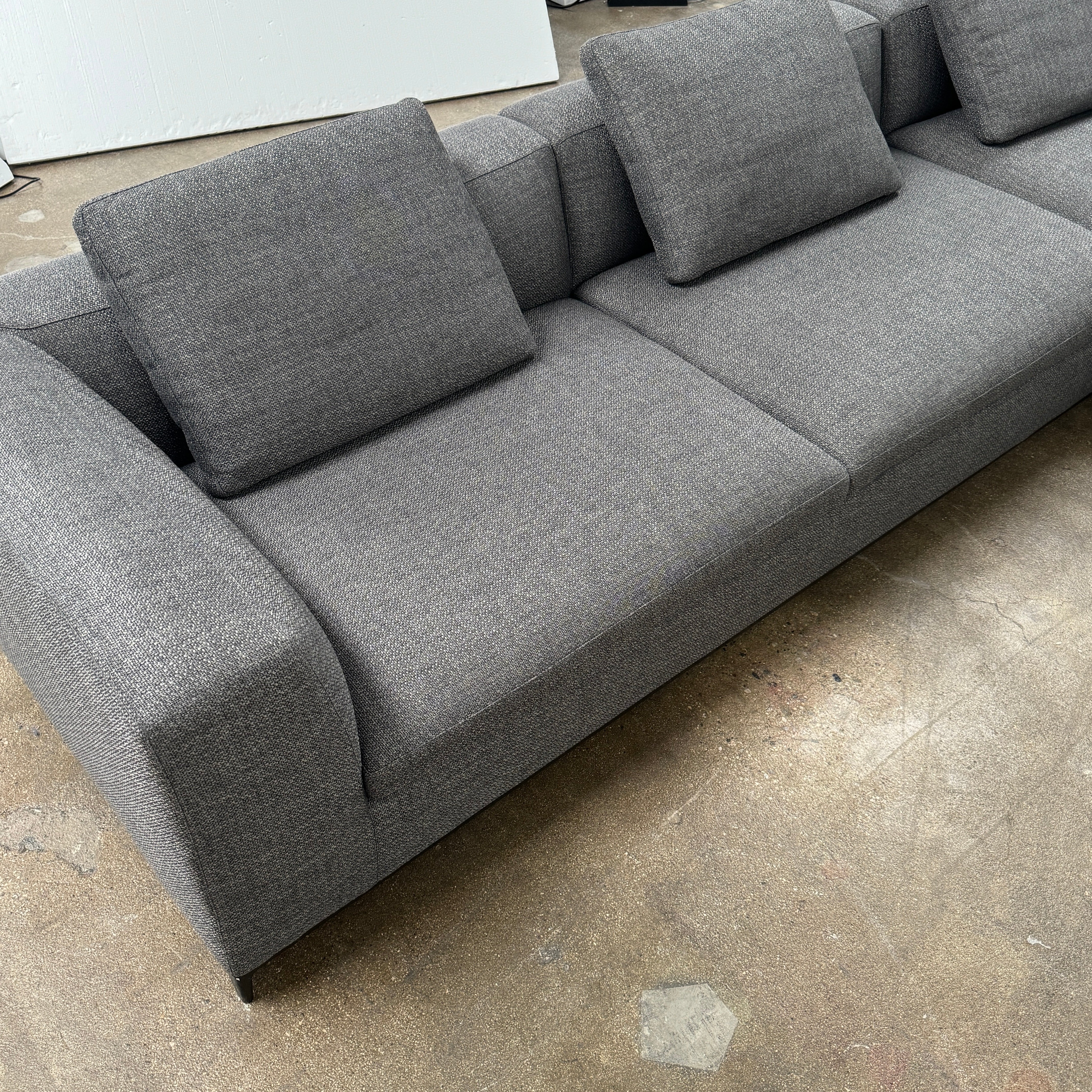 The B&B Italia Michele Club Sectional Sofa by B&B Italia, in dark grey open weave fabric with three seats and a right-side chaise lounge, sits on a concrete floor against a white wall—perfect for modern living spaces.