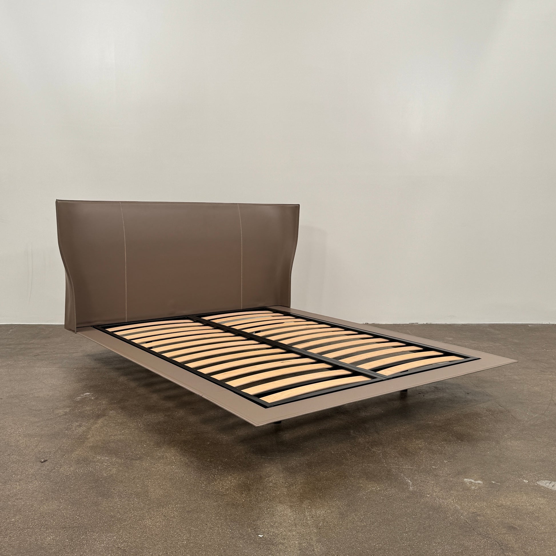 The B&B Italia Alys Queen Size Bed in Tortora Leather features a tall upholstered headboard and wooden slats, shown from a side angle against a plain background.
