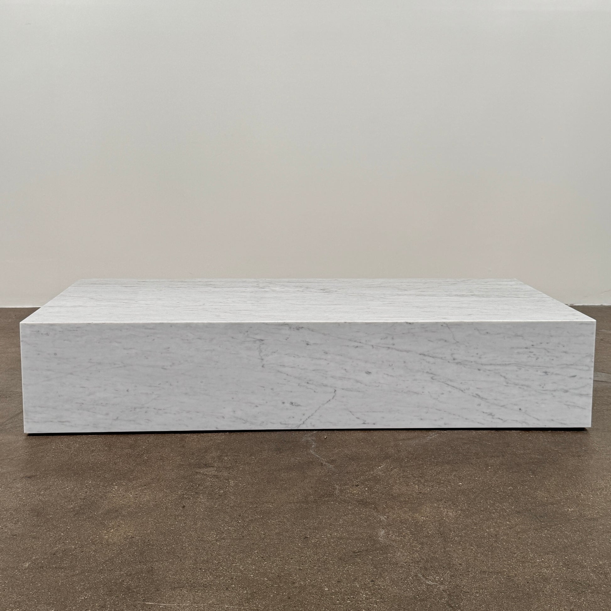 A minimalist room with light wood floors, an Audo Copenhagen Marble Plinth Table XL on a beige rug, a potted plant, and large windows draped with sheer white curtains.