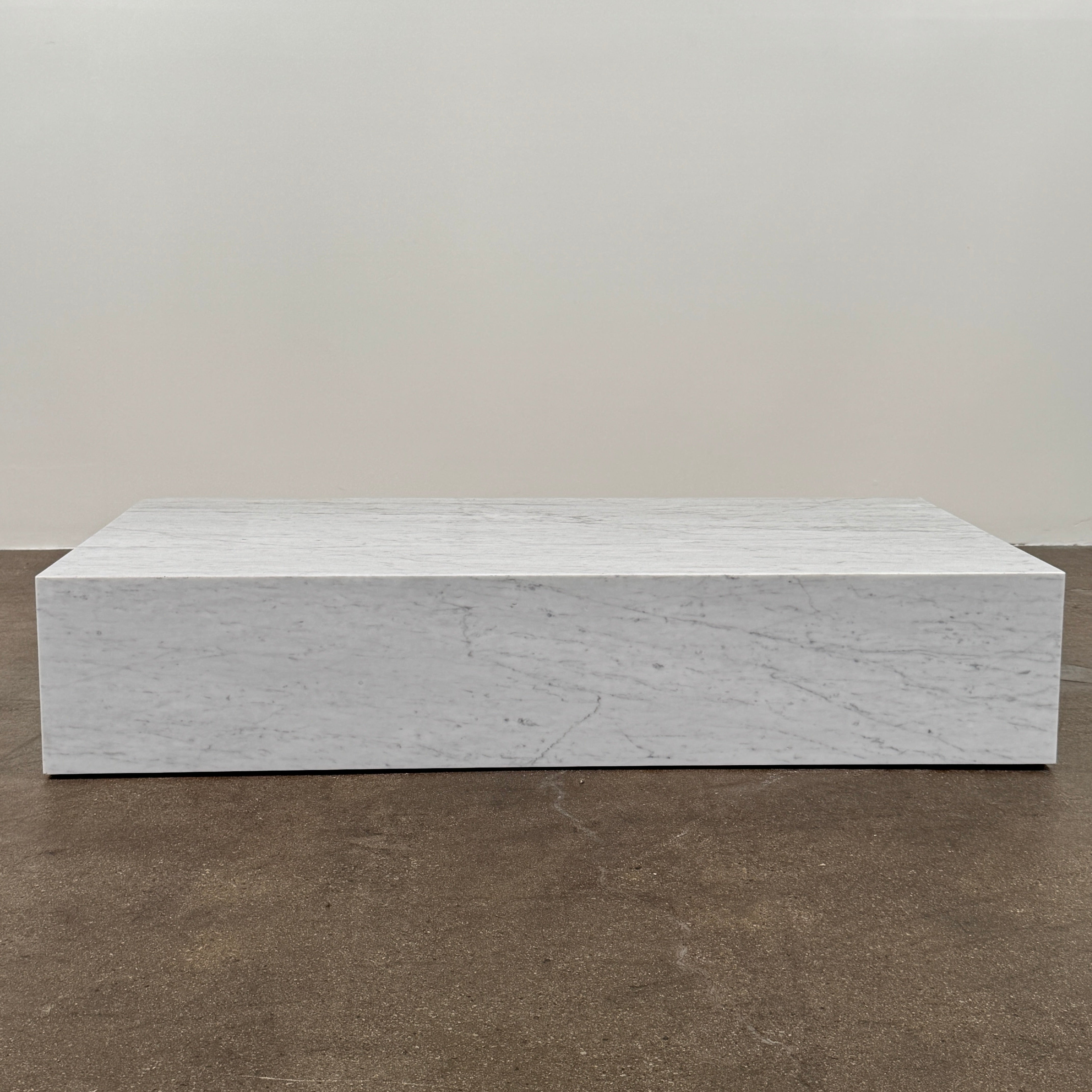 A minimalist room with light wood floors, an Audo Copenhagen Marble Plinth Table XL on a beige rug, a potted plant, and large windows draped with sheer white curtains.