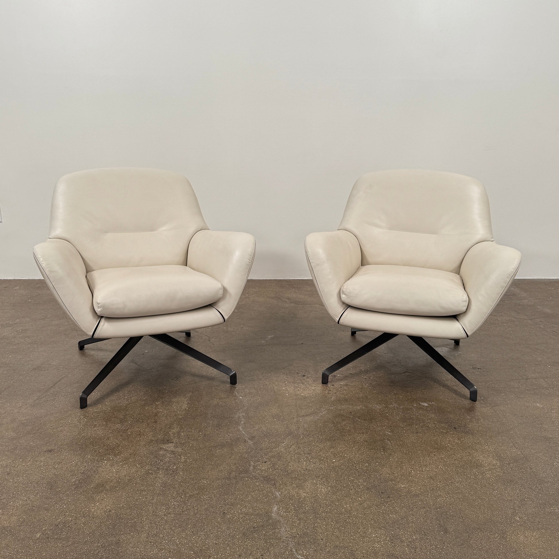 Two modern Minotti Jensen Swivel Chairs by Minotti face each other in a bright room with large windows overlooking the city, pale wood floors, and minimal decor for a contemporary, sophisticated look.