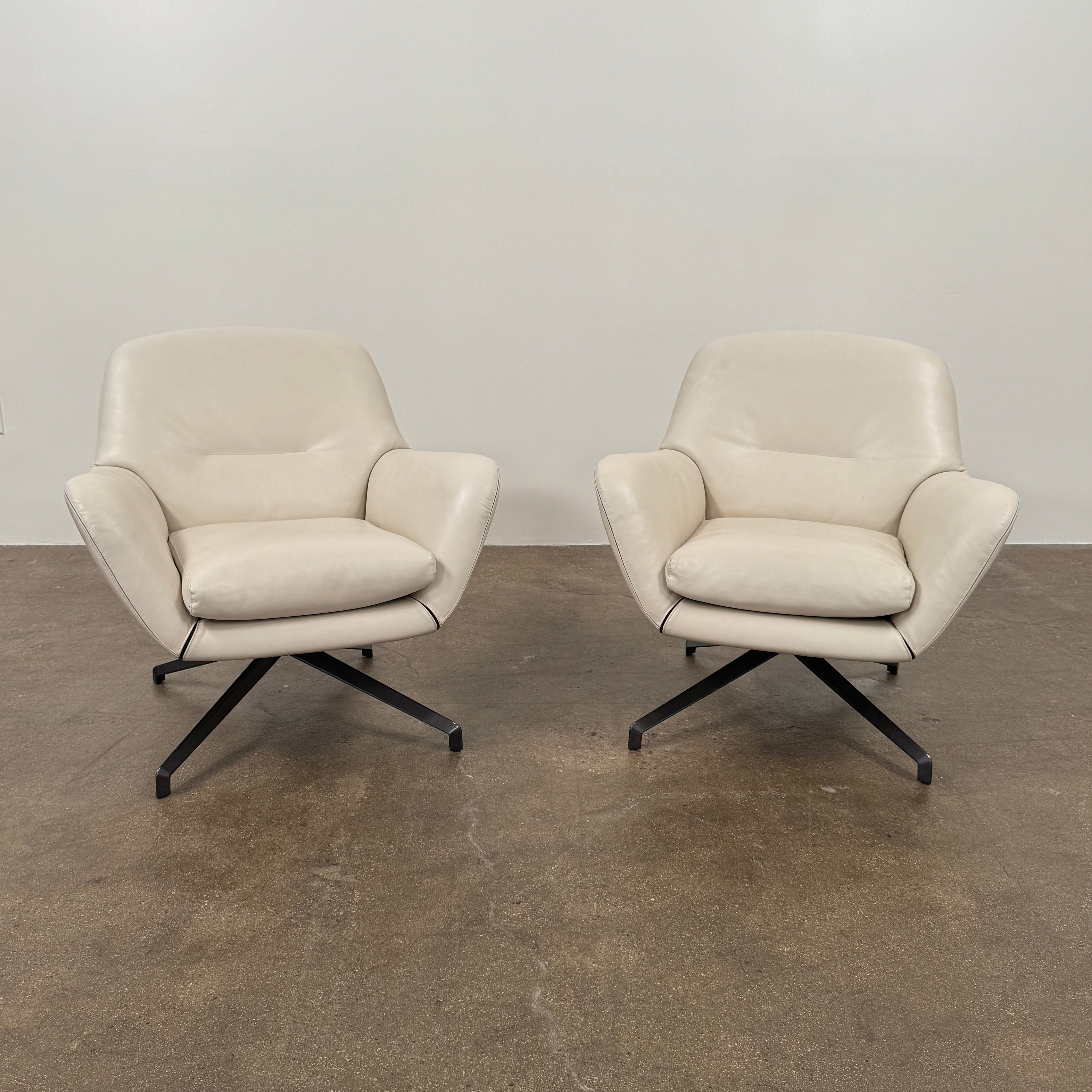 Two modern Minotti Jensen Swivel Chairs by Minotti face each other in a bright room with large windows overlooking the city, pale wood floors, and minimal decor for a contemporary, sophisticated look.