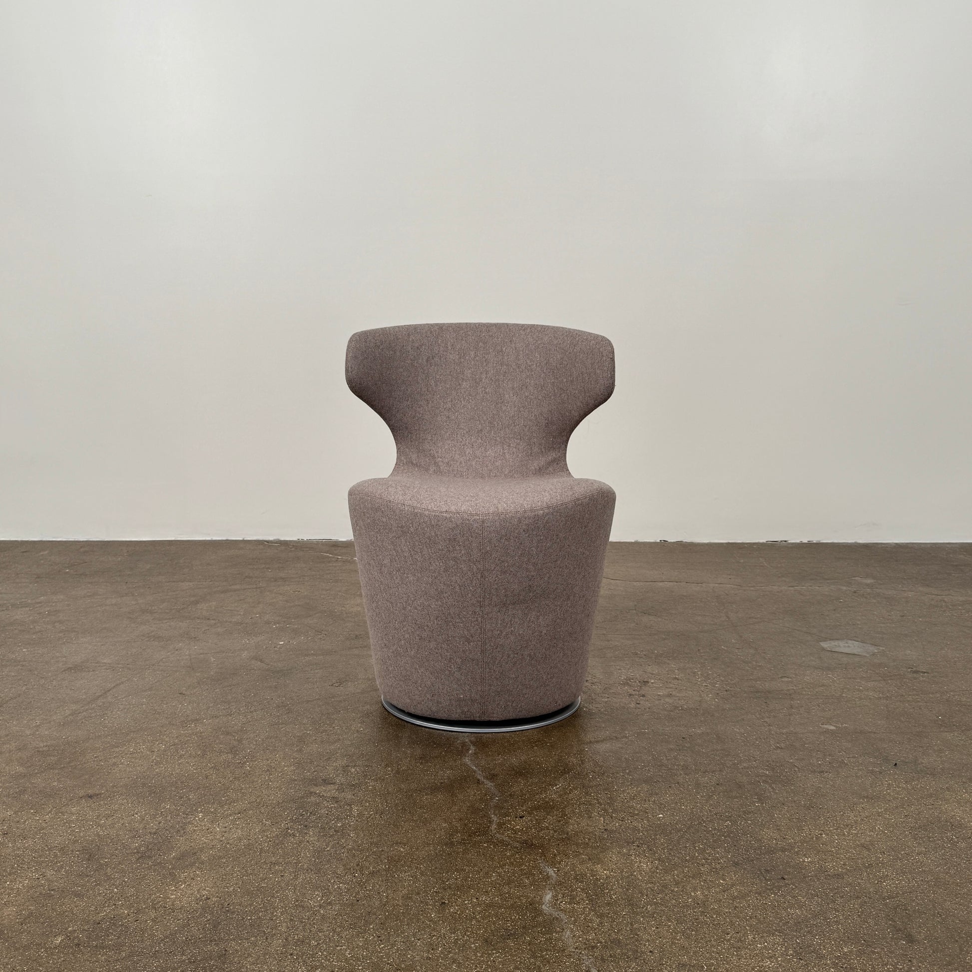 The B&B Italia Mini Papilio Swivel Chair features curved armrests and light brown upholstery, elegantly set on a polished concrete floor against a plain white wall.
