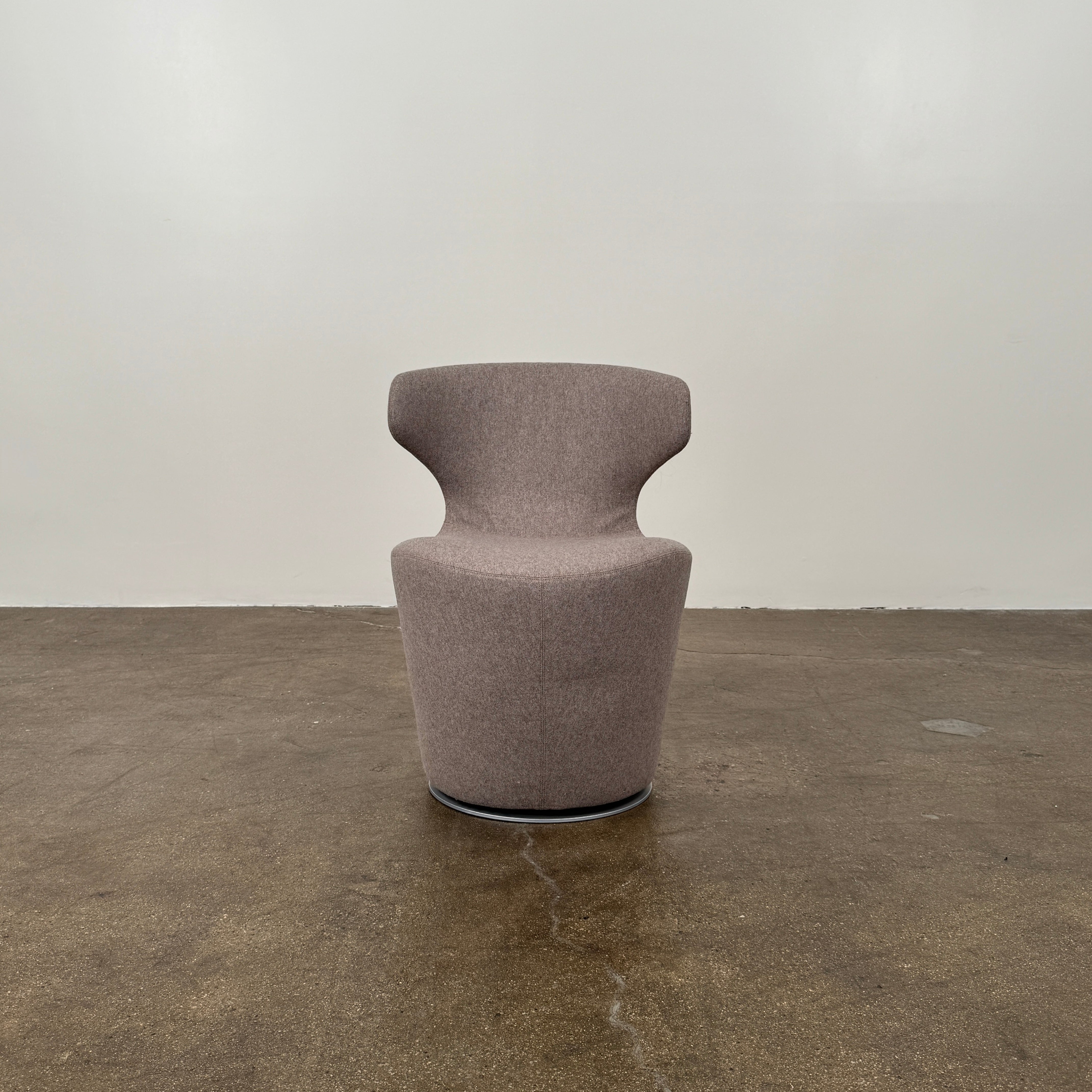 The B&B Italia Mini Papilio Swivel Chair features curved armrests and light brown upholstery, elegantly set on a polished concrete floor against a plain white wall.