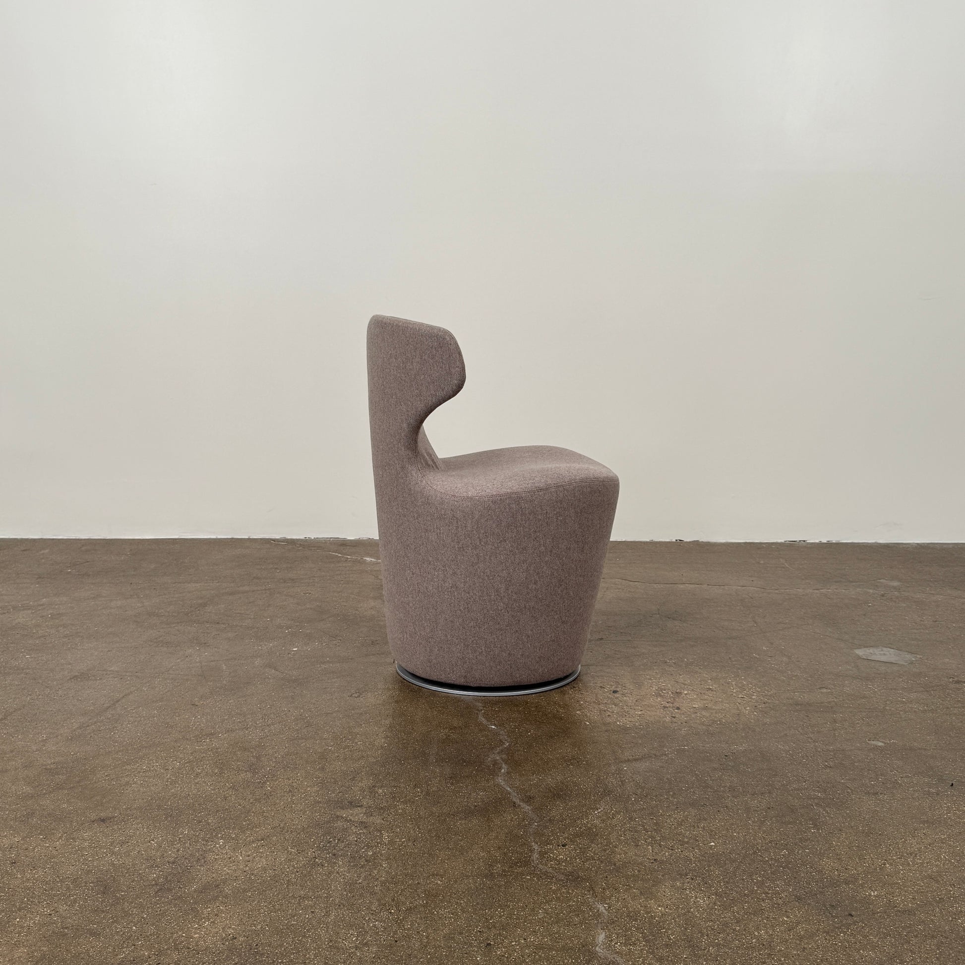 The B&B Italia Mini Papilio Swivel Chair features curved armrests and light brown upholstery, elegantly set on a polished concrete floor against a plain white wall.