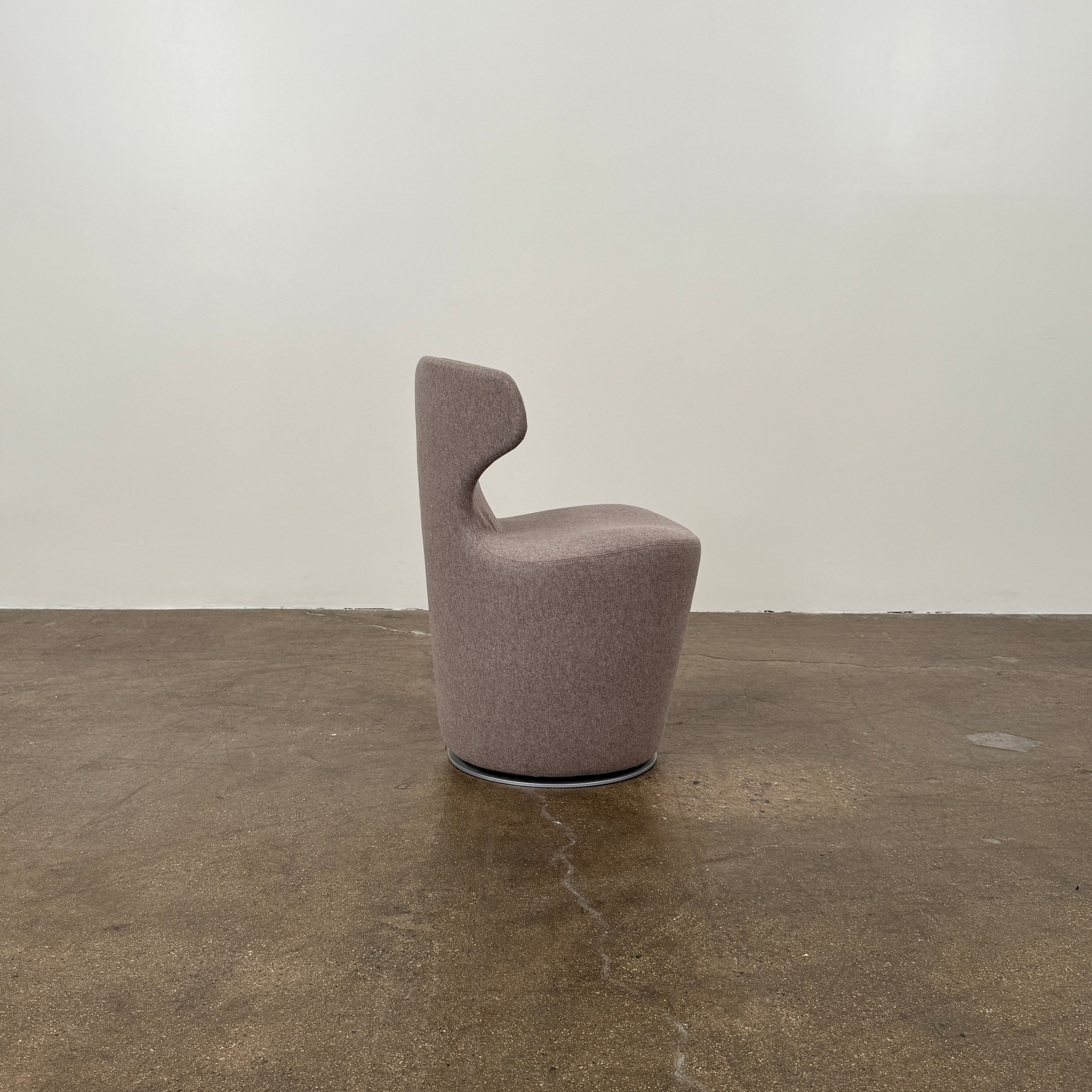 The B&B Italia Mini Papilio Swivel Chair features curved armrests and light brown upholstery, elegantly set on a polished concrete floor against a plain white wall.