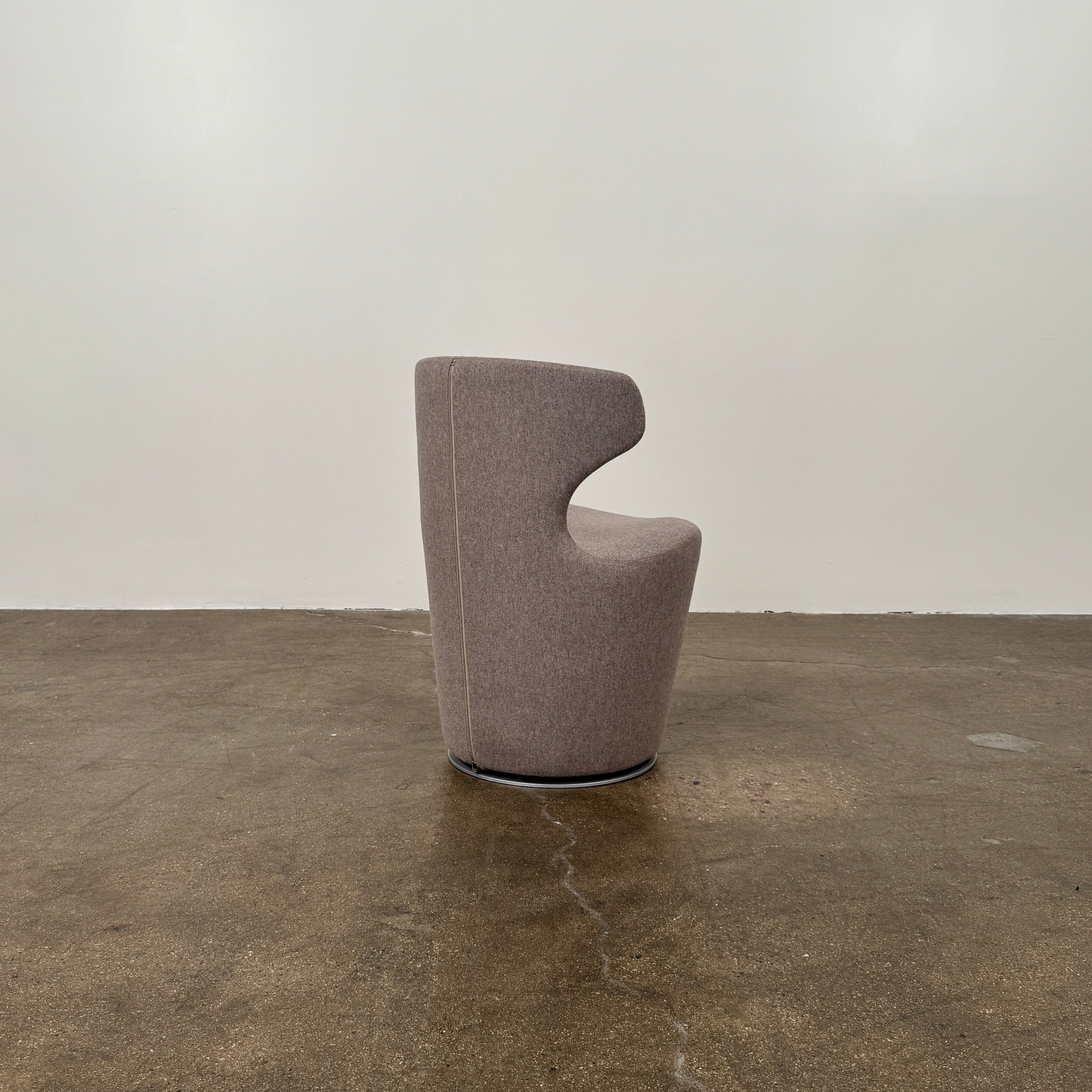 The B&B Italia Mini Papilio Swivel Chair features curved armrests and light brown upholstery, elegantly set on a polished concrete floor against a plain white wall.