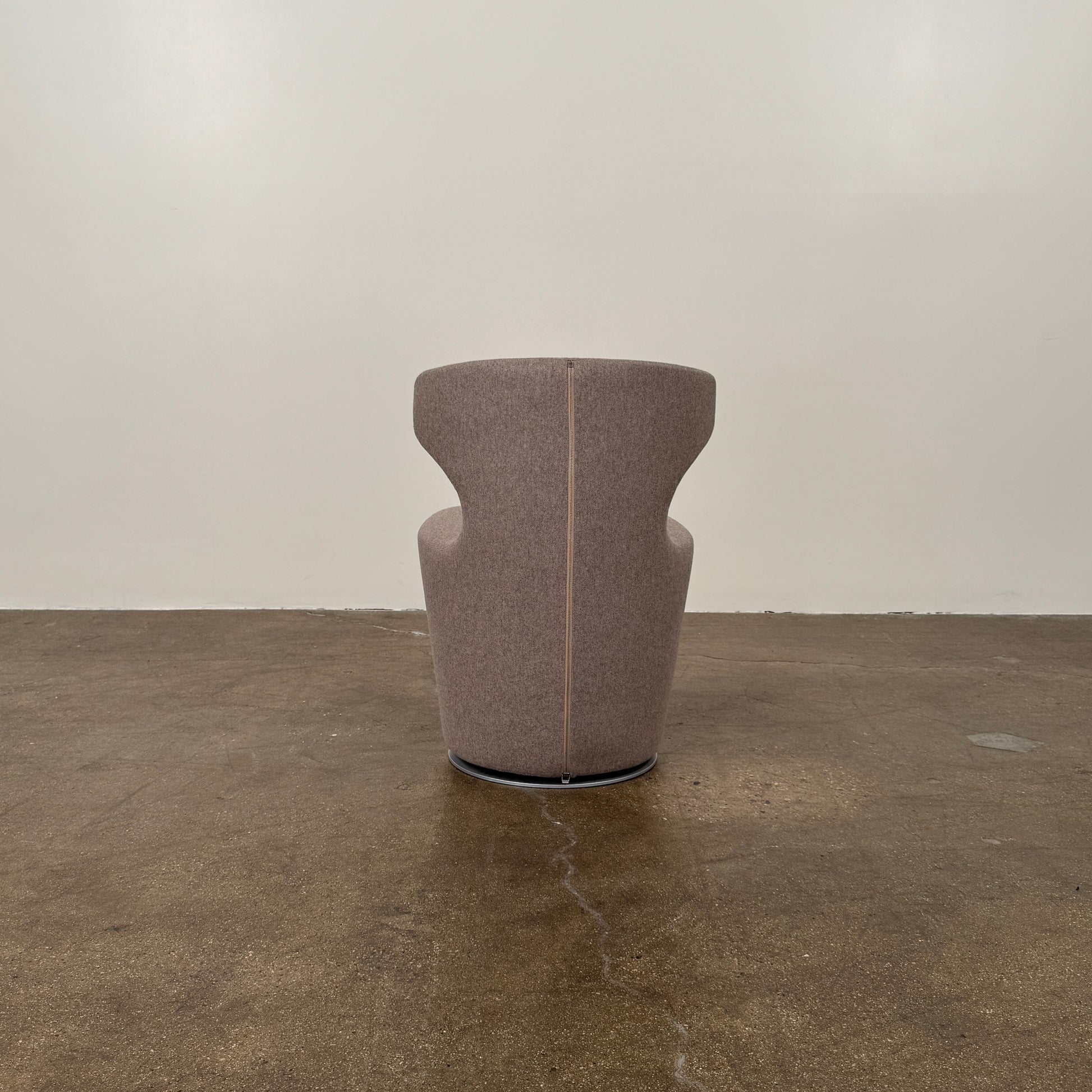The B&B Italia Mini Papilio Swivel Chair features curved armrests and light brown upholstery, elegantly set on a polished concrete floor against a plain white wall.