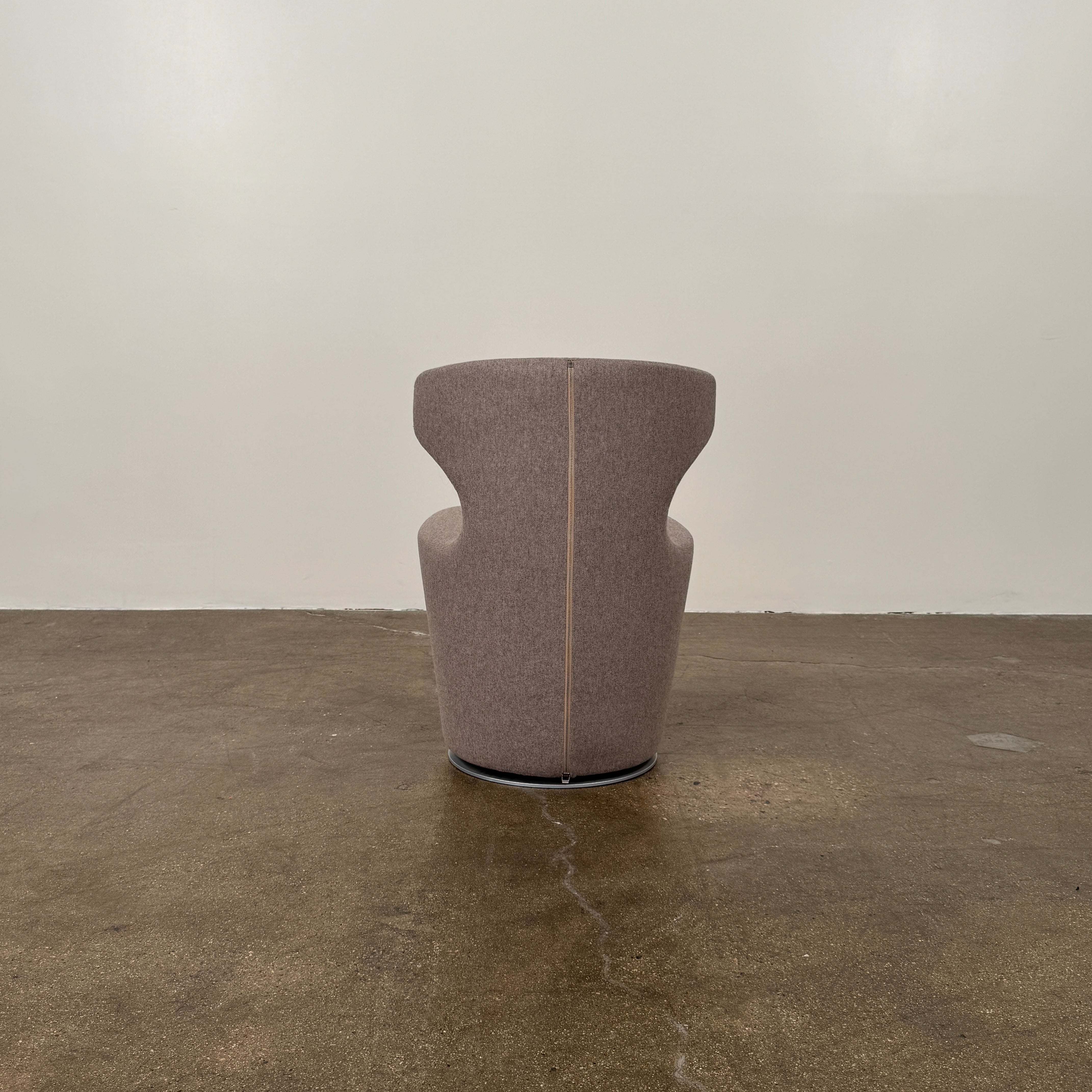 The B&B Italia Mini Papilio Swivel Chair features curved armrests and light brown upholstery, elegantly set on a polished concrete floor against a plain white wall.