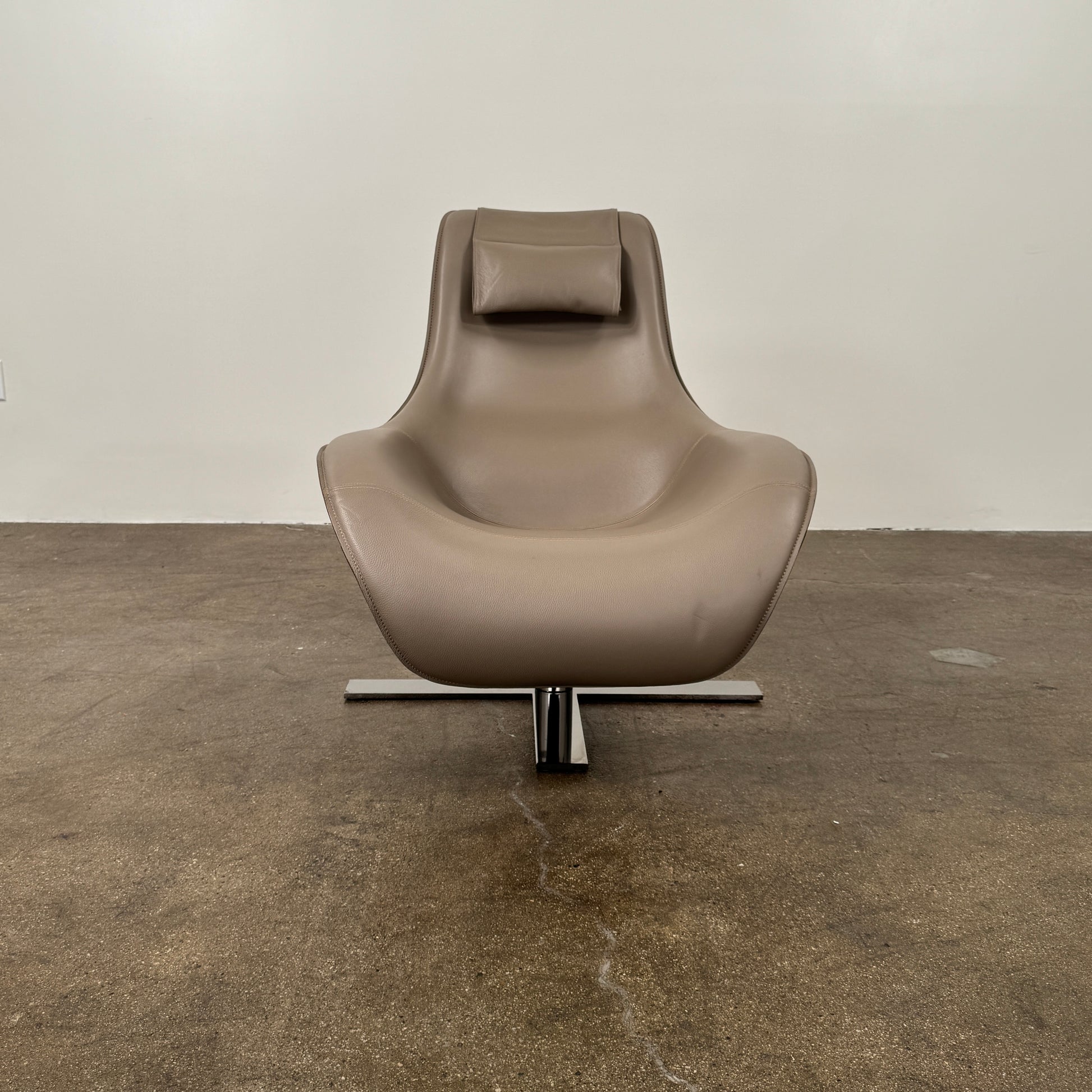 The B&B Italia Mart Armchair with Swivel Base is a modern beige lounge chair featuring a low backrest and headrest, set on a sleek swivel base against a plain white wall and concrete floor.